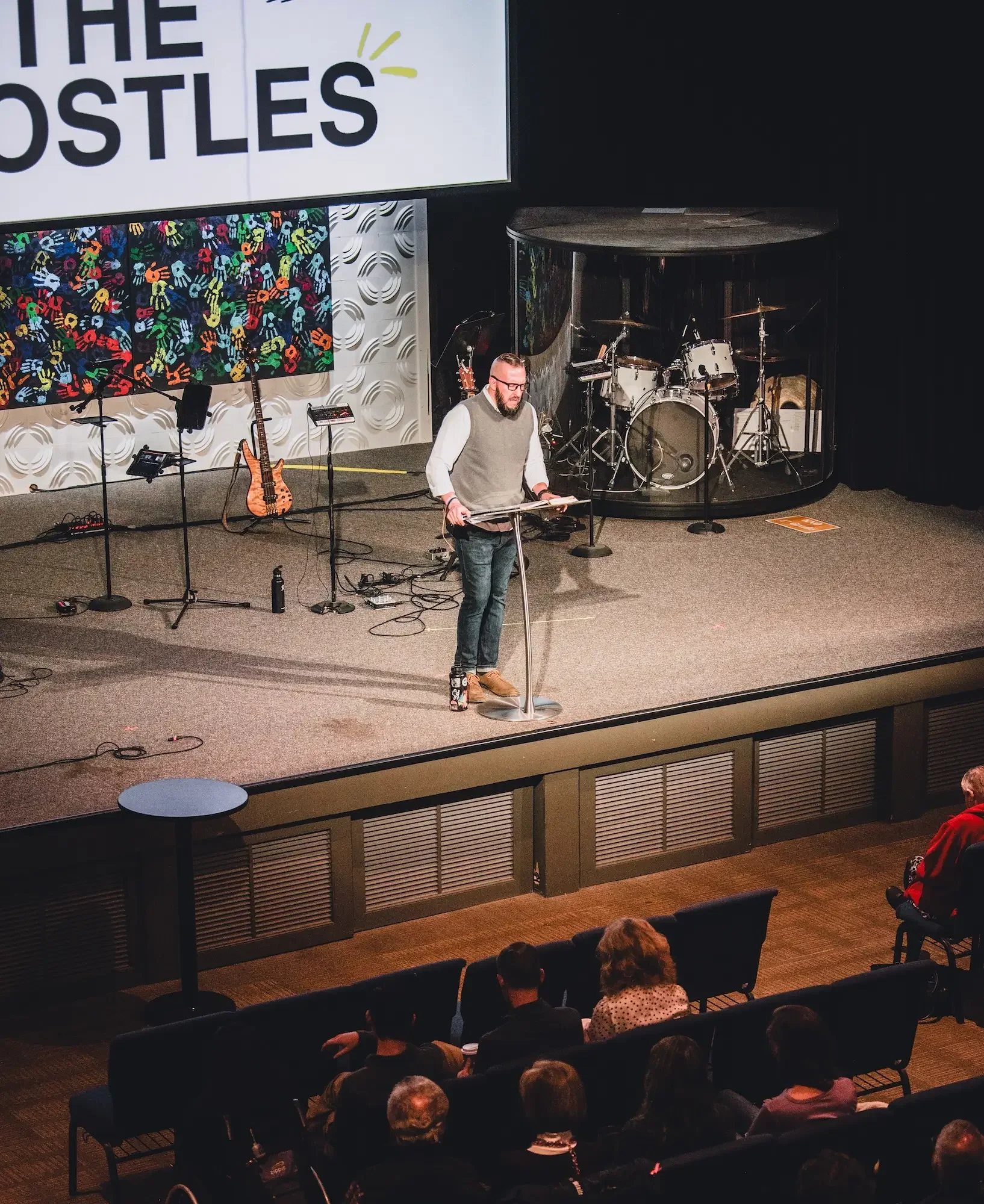 A man standing at a lectern on a stage, reading from a book. Behind him are musical instruments including a drum set and guitars. An audience is seated in front of the stage, attentively listening.