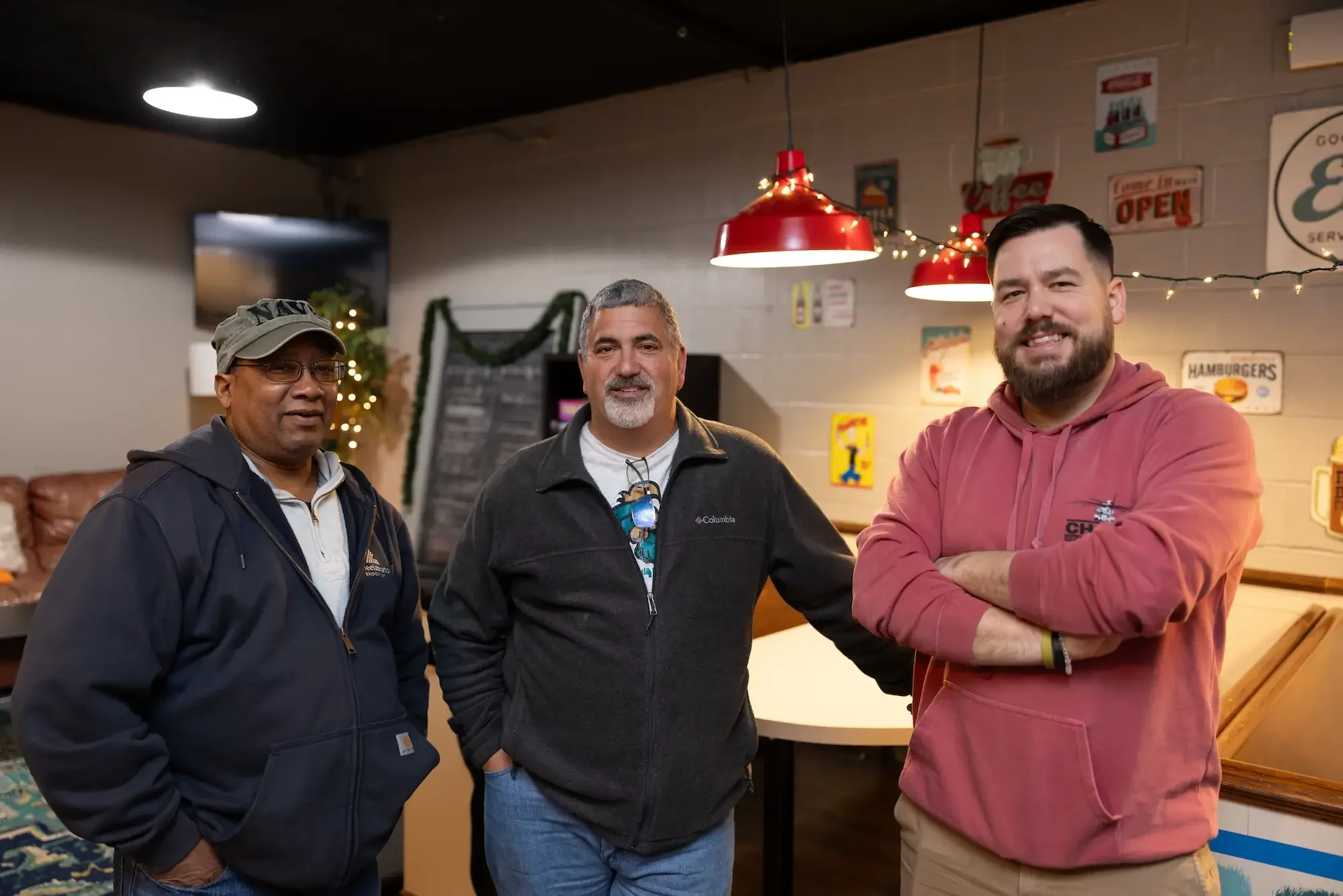 Three men inside a restaurant, standing in front of a counter, with decorations and signs on the wall behind them. They are smiling and posing for the photo.