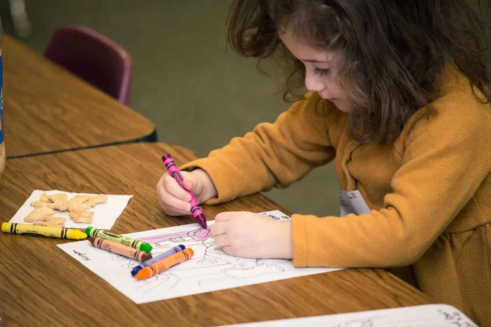 A young girl with curly brown hair, wearing an orange sweatshirt, is sitting at a wooden table coloring a drawing with a pink crayon. There are other crayons and cookies on a napkin nearby.