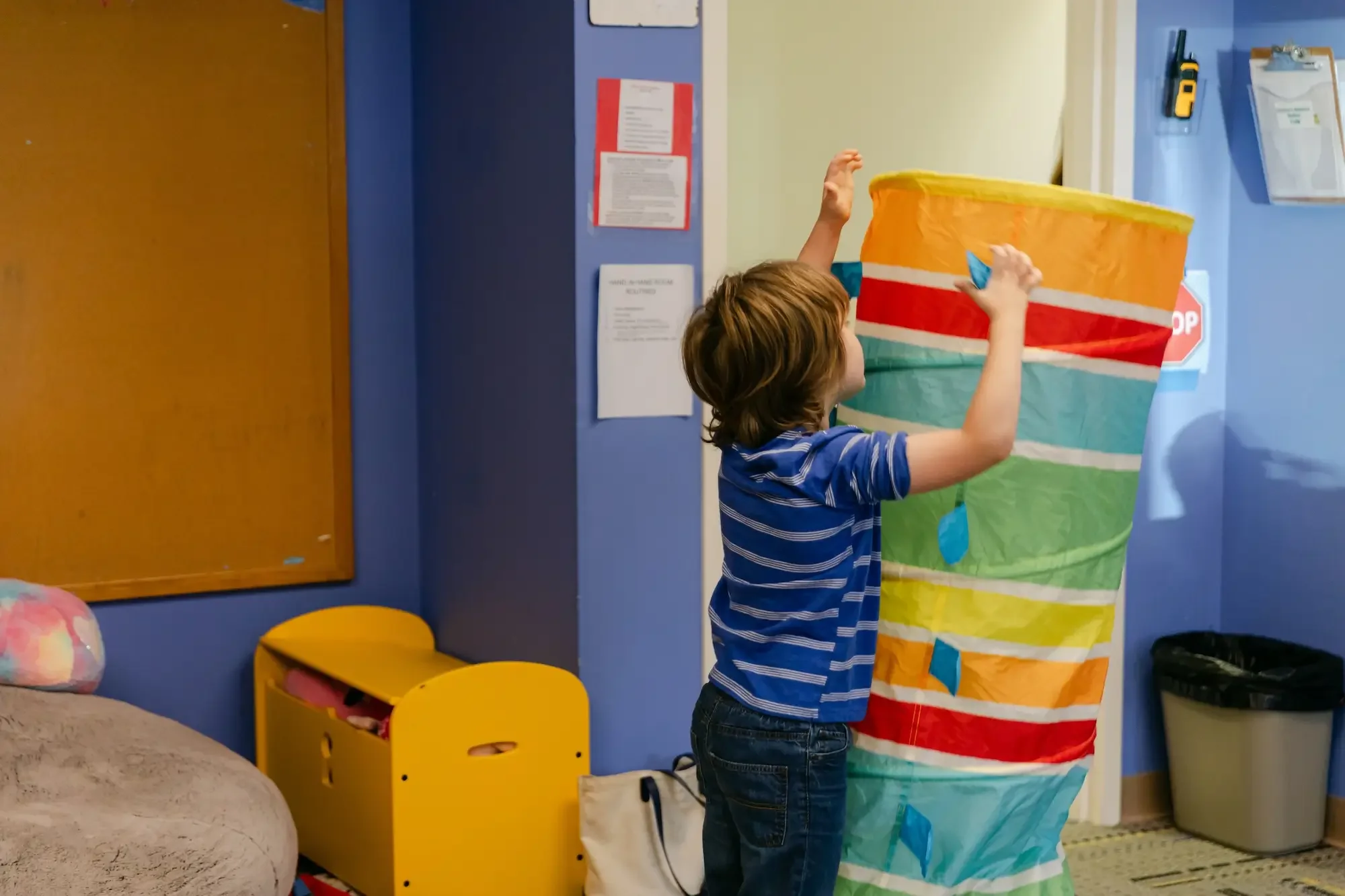 A young boy playing with a large colorful collapsible tube in a room with blue walls and a bulletin board.