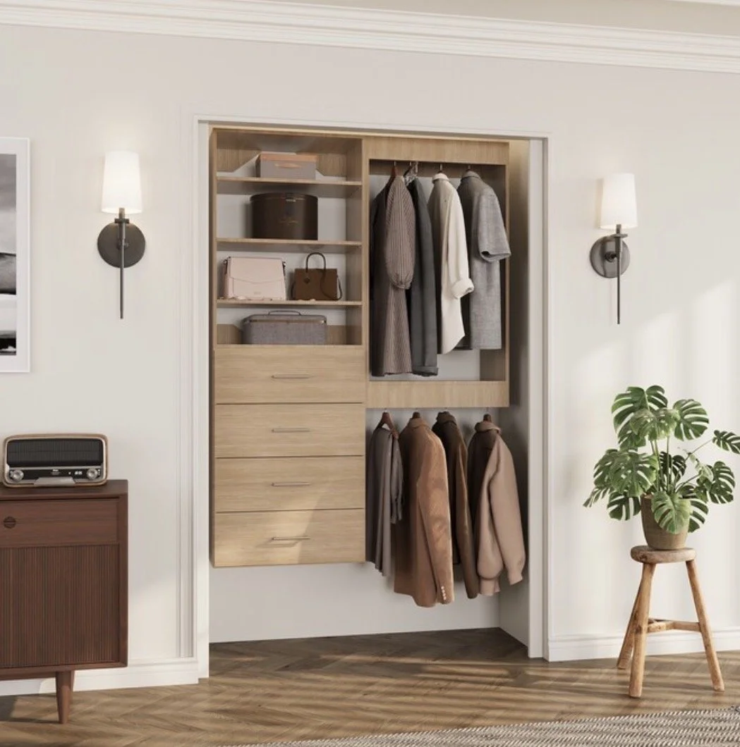 An open closet with wood shelving and hanging space, holding clothes and bags, in a room with wall sconces and a potted plant.