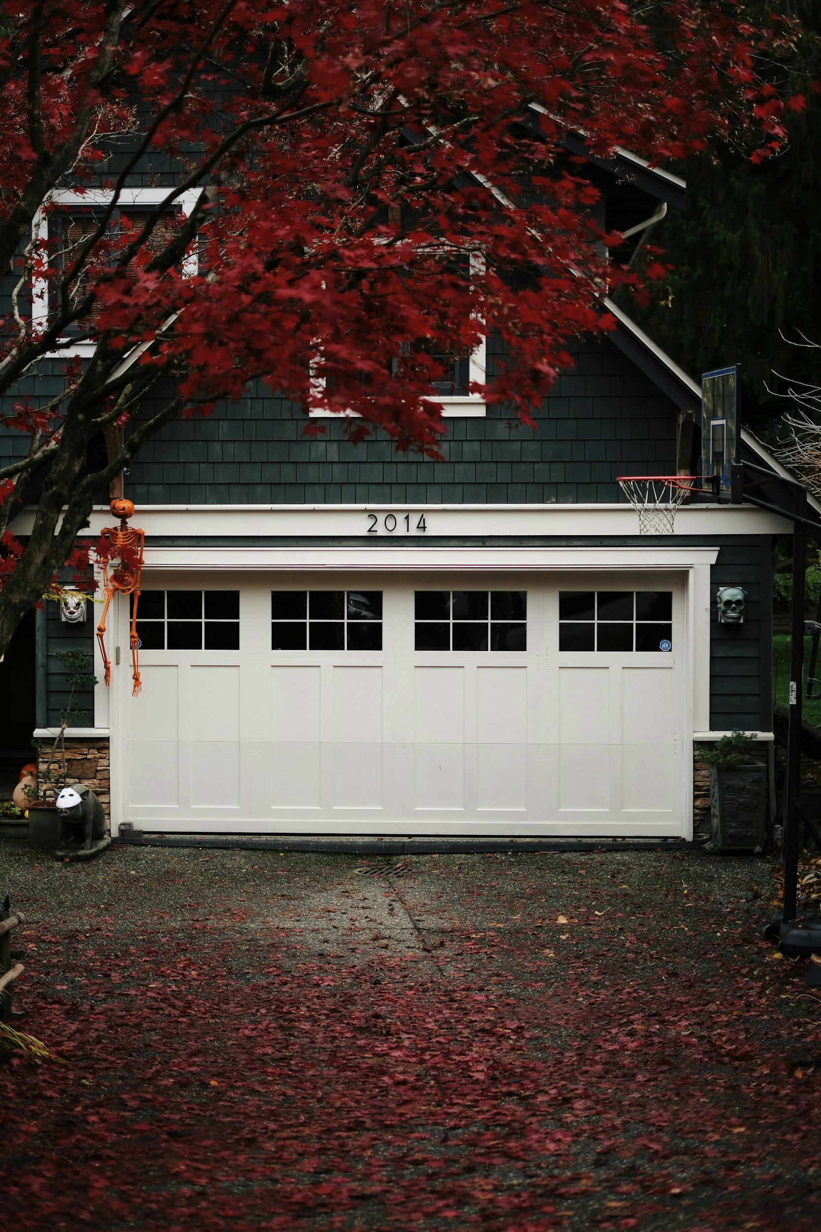 A house with a dark green exterior, white garage door, and the year 2014 above the garage. A tree with red leaves partially covers the house, and autumn leaves are fallen on the ground.