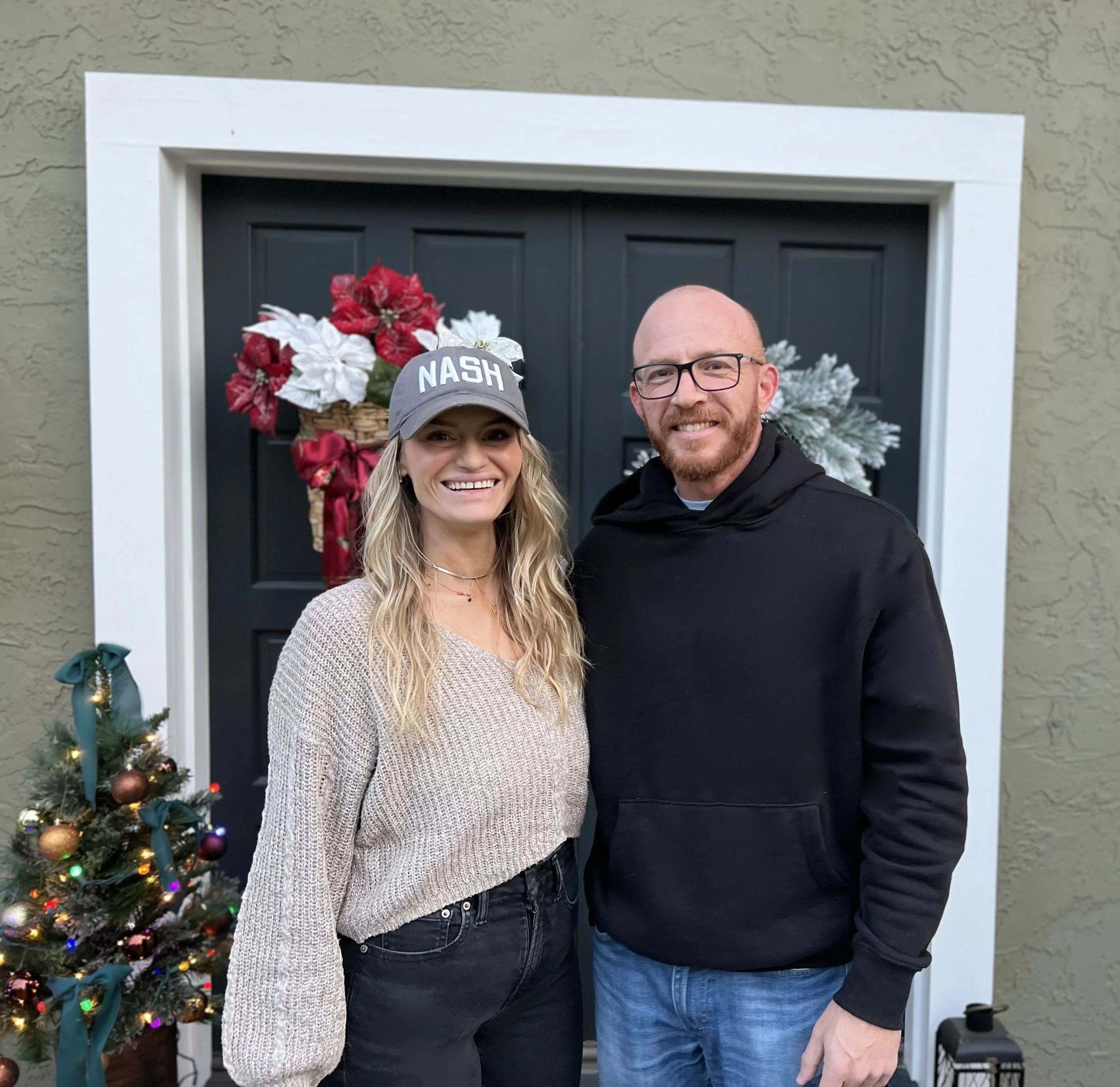 A woman and man standing in front of a decorated door with holiday decorations, smiling at the camera.