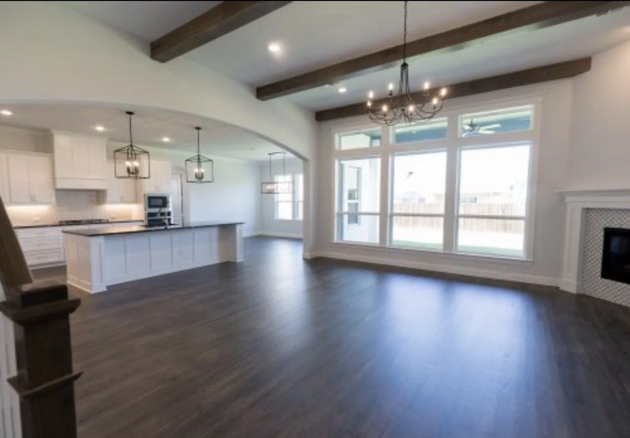 Empty living room and kitchen area with large windows, dark wood flooring, white walls, and wooden ceiling beams, featuring pendant light fixtures and a fireplace.