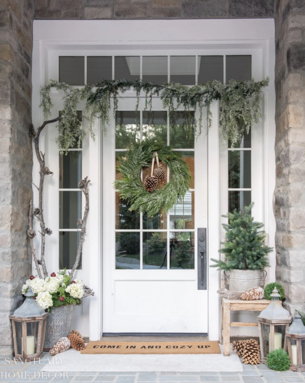 Decorated front porch with a white door, green wreath with pinecones, greenery garland above the door, small potted evergreen tree and flower arrangement on the right, lanterns and pinecones on the ground, doormat with the words 'Come in and cozy up'.