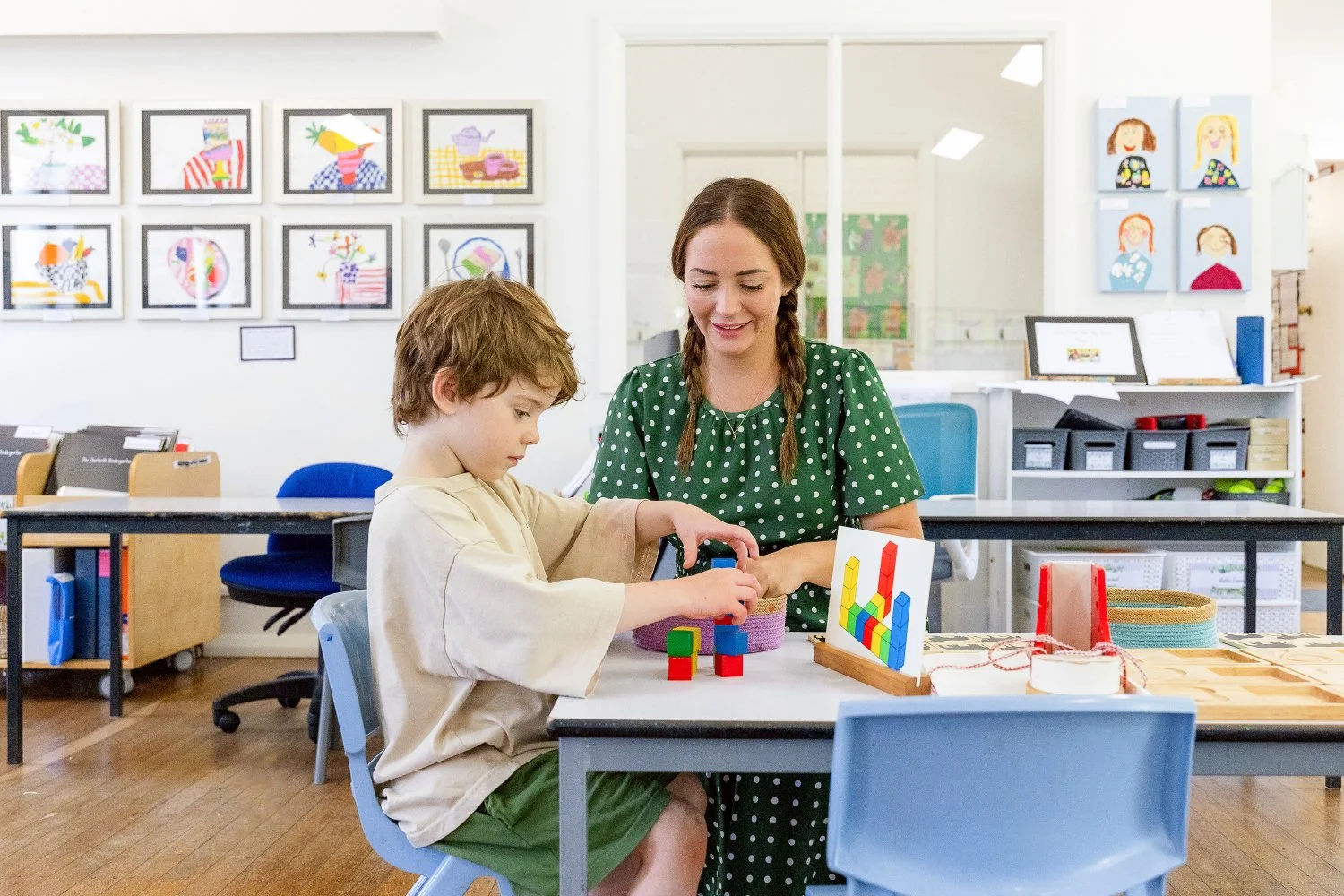 A young boy and a woman, possibly a teacher or caregiver, sitting at a table in a classroom, playing with colorful building blocks.
