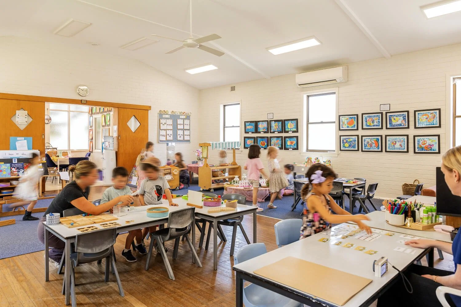 Children and teachers in a colorful classroom engaged in arts and crafts activities, with artwork displayed on the walls and tables with art supplies.