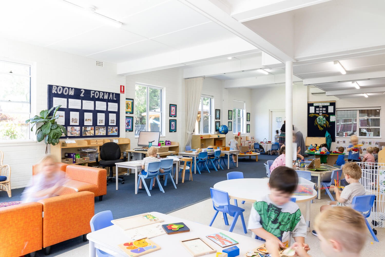Children playing and learning in a bright, colorful classroom with educational displays, tables, and chairs.
