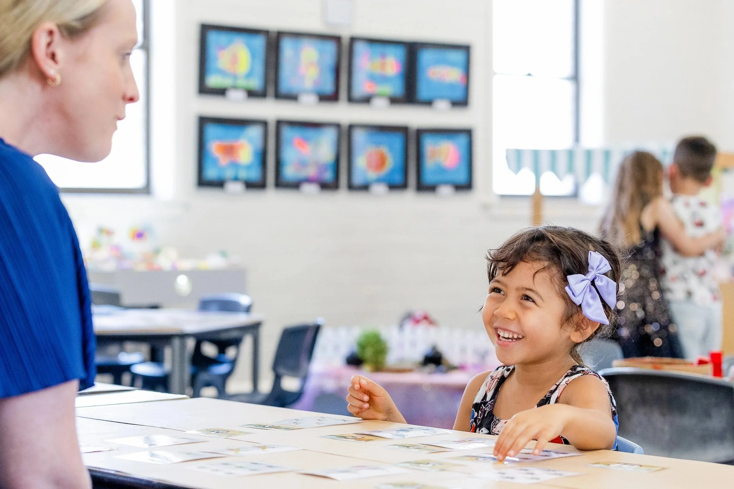 A young girl with a purple bow in her hair smiling and interacting with an adult woman, sitting at a table in a classroom or activity center with artwork on the walls.