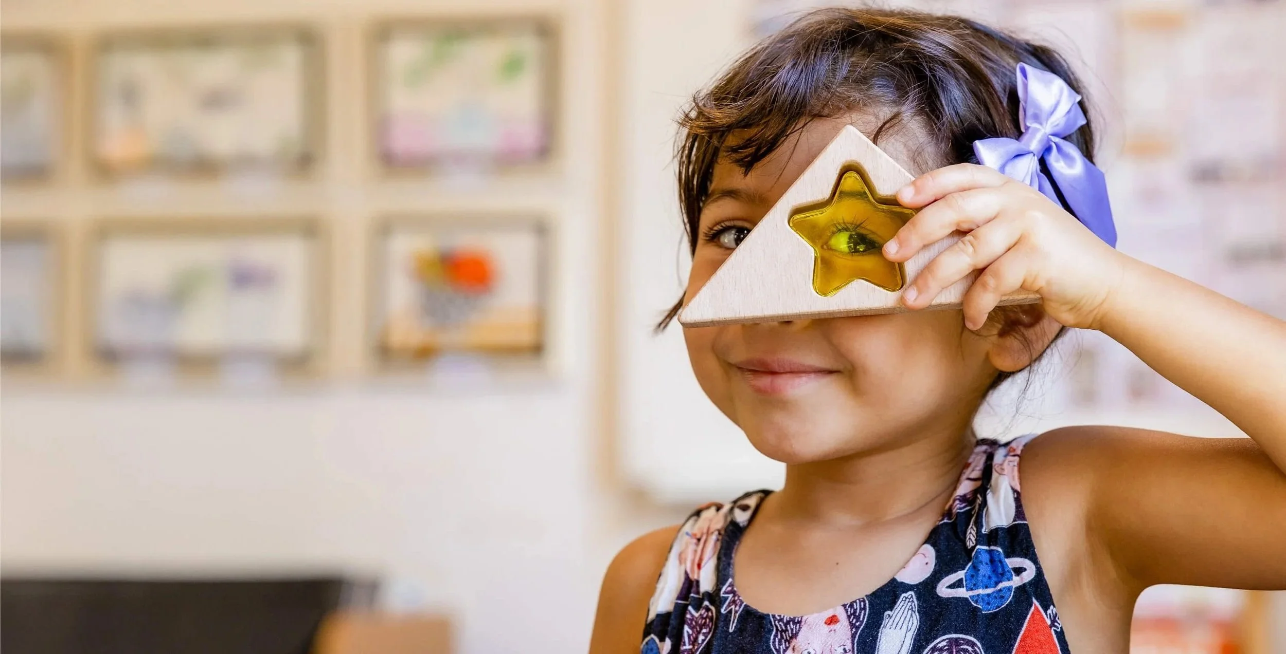 A young girl with dark hair and a purple bow in her hair, smiling while holding a wooden shape with a star-shaped hole up to her eye, in a bright room.