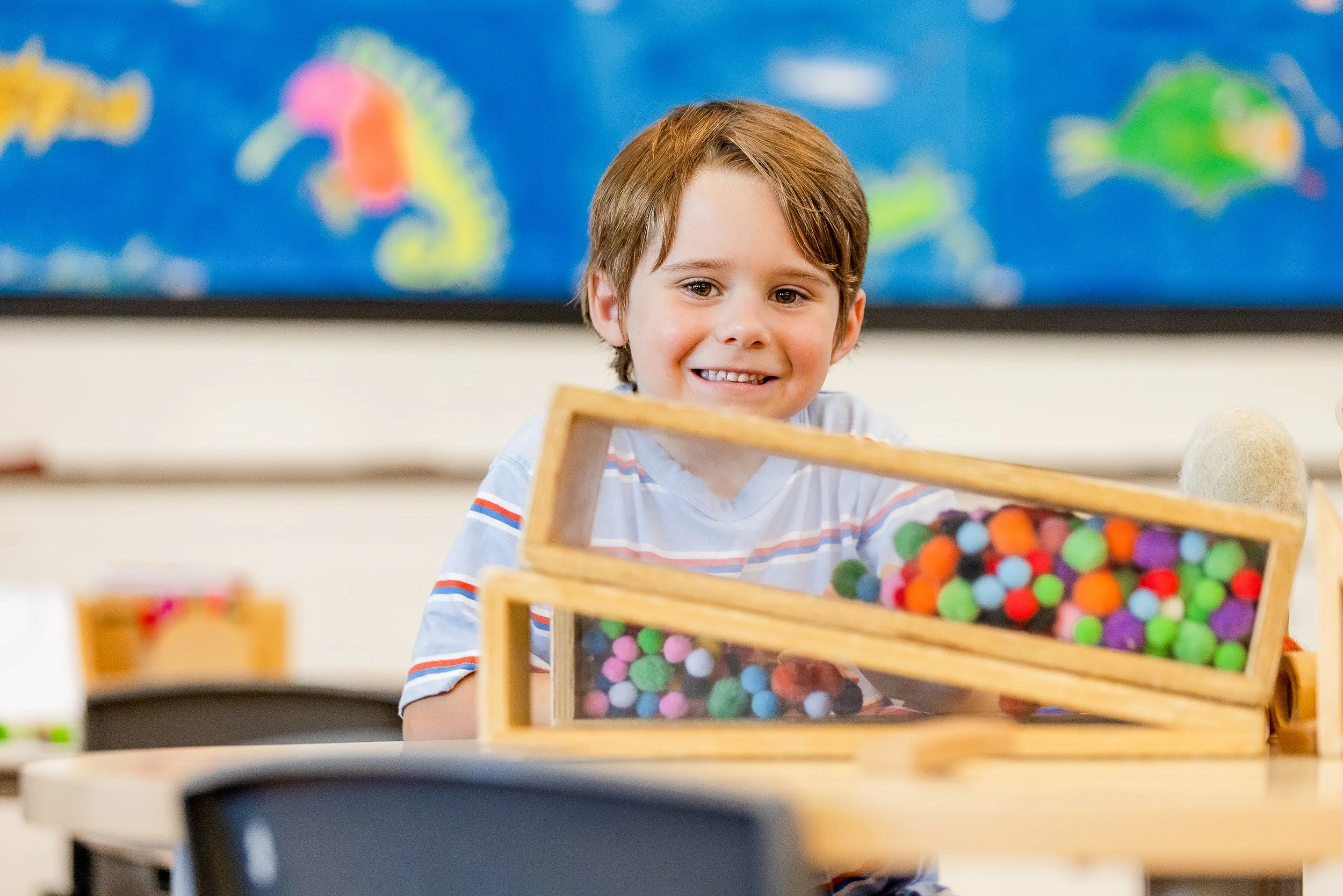 A young boy with brown hair smiling at a wooden box filled with colorful pom-poms in a classroom setting.