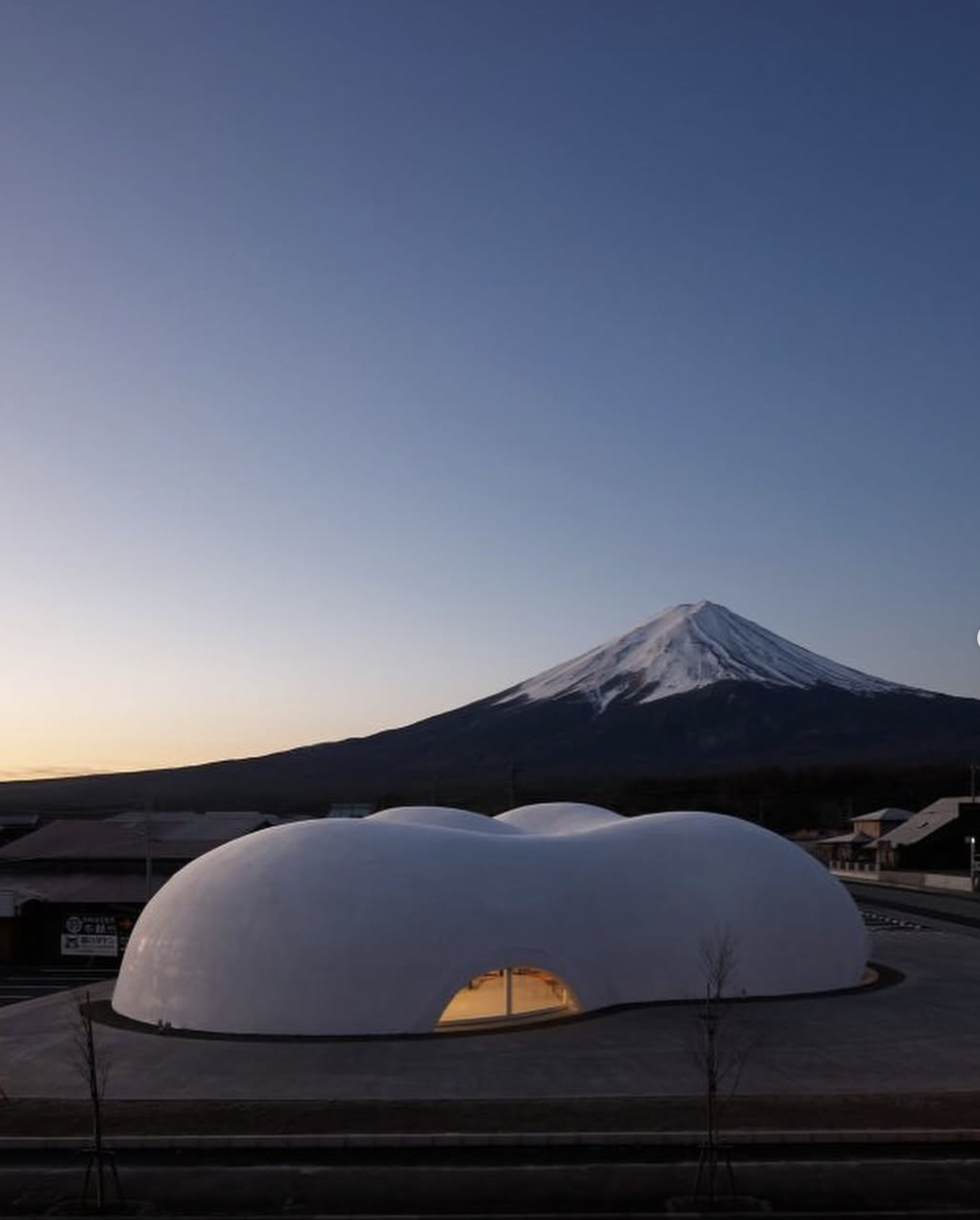 View of Mount Fuji with a snow-capped peak in the background and a modern, dome-shaped building with glowing interior lights in the foreground.