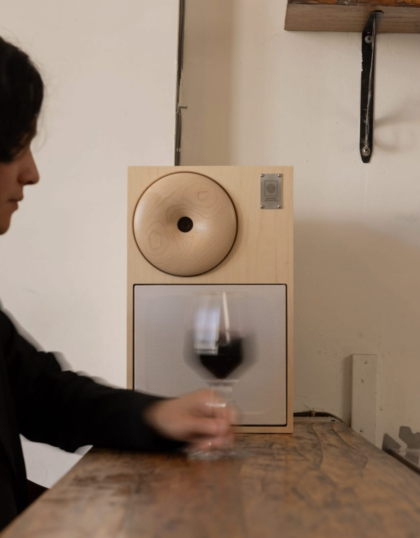 A person holding a glass of red wine in front of a wooden speaker on a table. The speaker has a large, round wooden dome on top and a rectangular speaker grille below. The background features a white wall with some mounted hardware and a small metal plate.