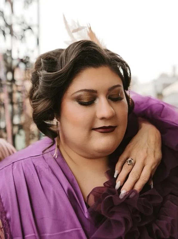 A woman with makeup and dark lipstick, wearing a purple gown with ruffles, and a white feathered headpiece, posing with her eyes closed and resting her hand on her chest.