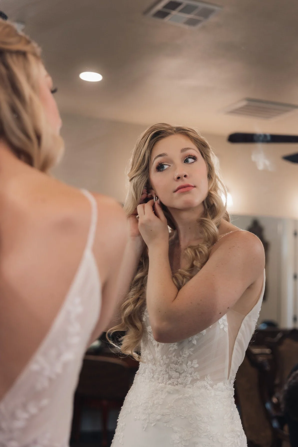 Young woman in a white wedding dress with lace details is looking at herself in a mirror while adjusting her earring in a room with warm lighting.