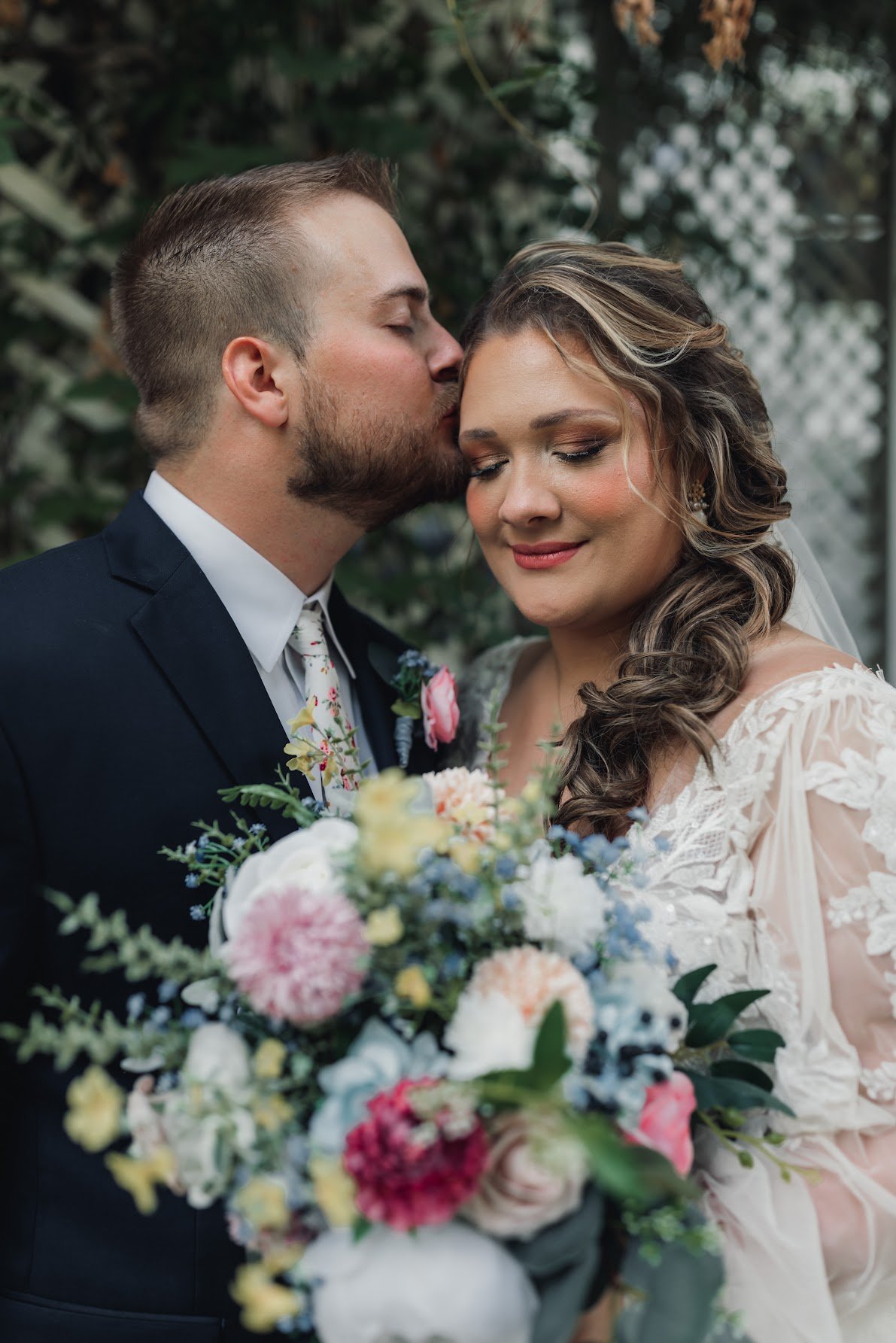 A groom kissing a bride on the forehead during their wedding, with a bouquet of colorful flowers, standing outdoors against a leafy background.