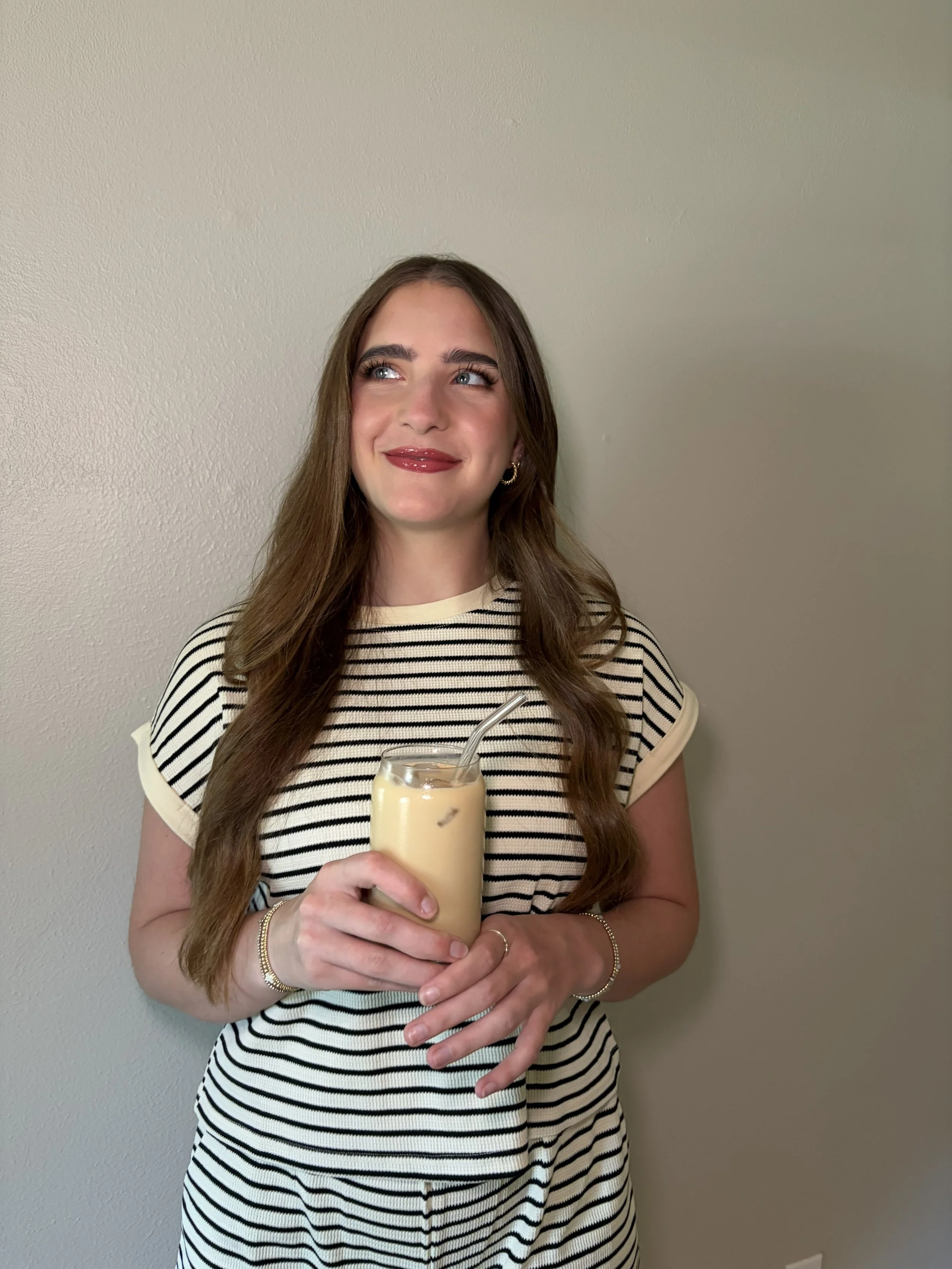 A young woman with long brown hair and blue eyes holding a glass of iced coffee or tea with a straw, wearing a black and white striped shirt and smiling while looking to the side, standing against a plain beige wall.