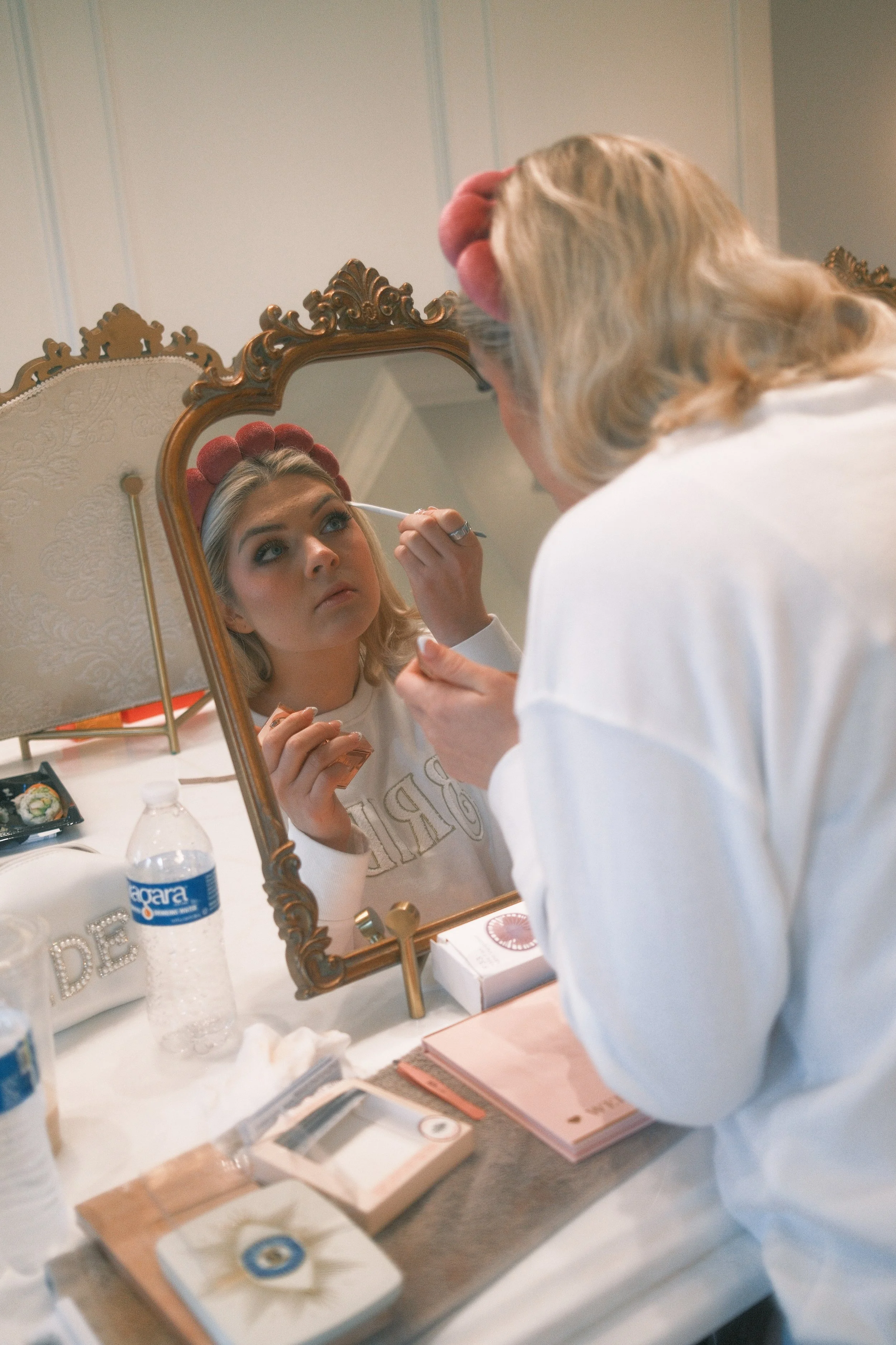 A young woman in a white shirt with a pink makeup headband, applying makeup to her eyebrows using a brush while looking into a vintage decorative mirror.
