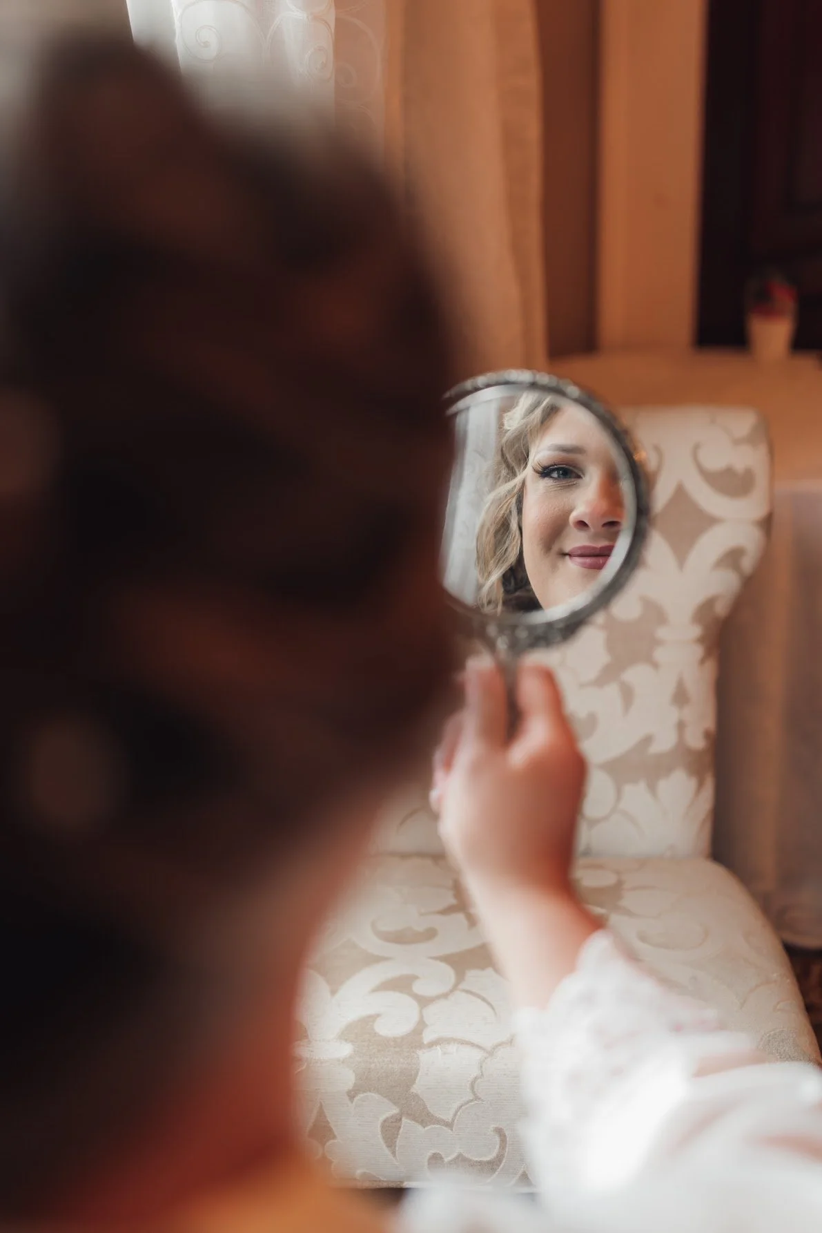 A woman holds a small mirror, viewing her reflection which shows her smiling face with styled hair and makeup.