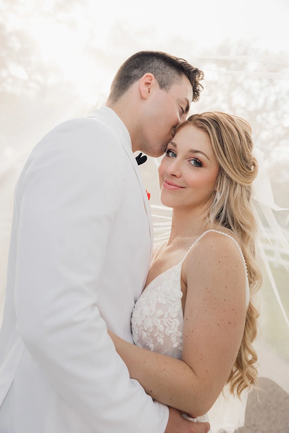 A bride and groom embrace on their wedding day, with the groom kissing the bride on her forehead and the bride smiling at the camera.