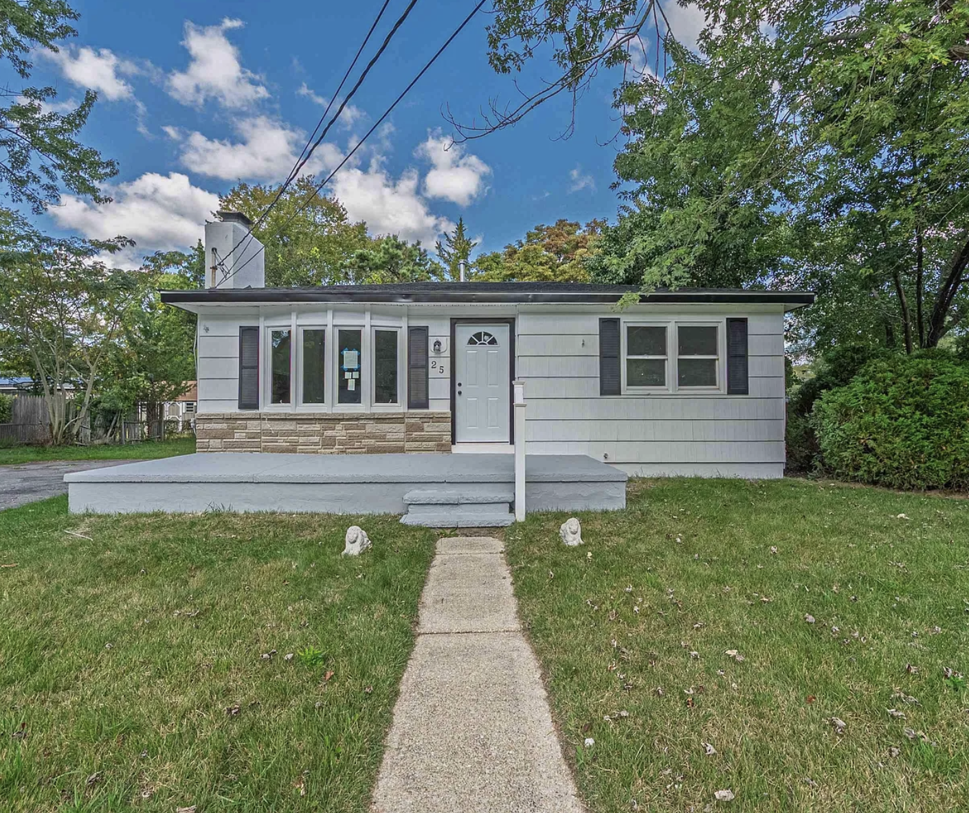Front view of a single-story white house with black shutters, a porch, and a concrete walkway, surrounded by a green lawn and trees under a partly cloudy sky.