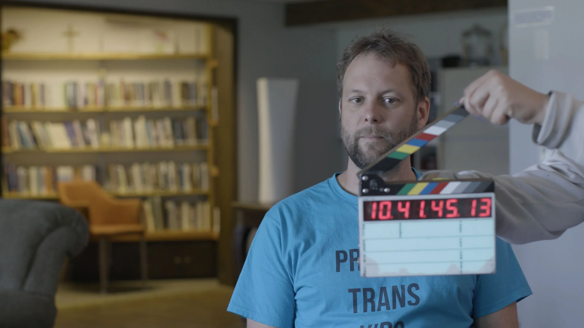 Man in blue T-shirt sitting in a room with bookshelves in the background, as a film crew member holds a clapboard near his face.