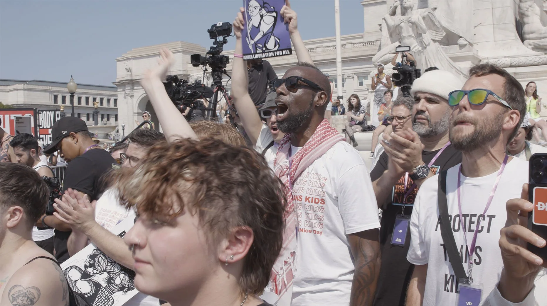 Crowd of people protesting outdoors on steps with statues, some clapping, holding signs, wearing sunglasses and casual clothing.
