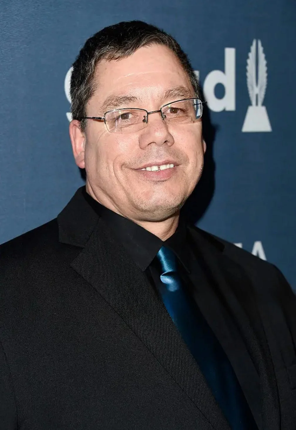 Man with glasses in a black suit and blue tie smiling at an event.