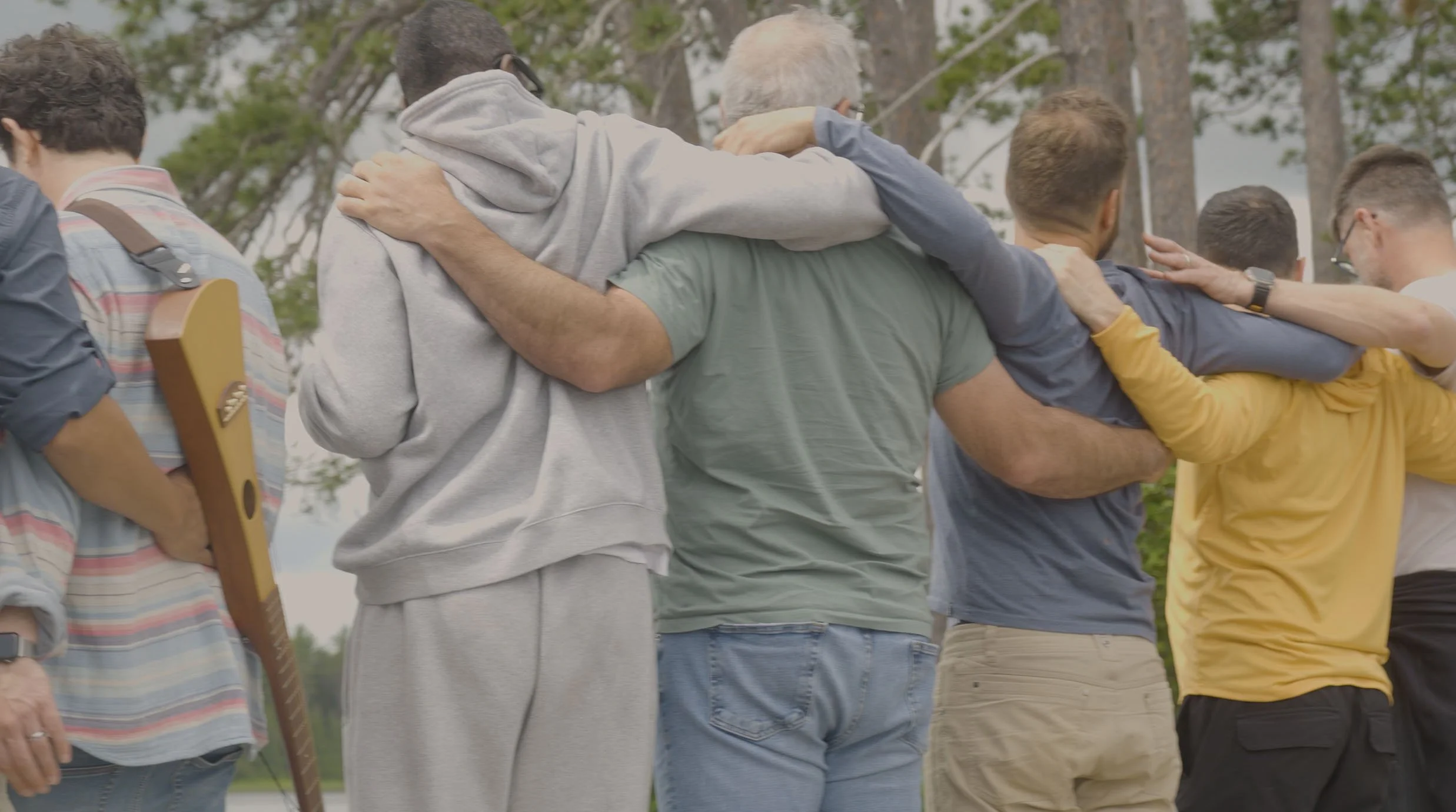 Group of people standing outdoors with arms around each other's shoulders in a circle.