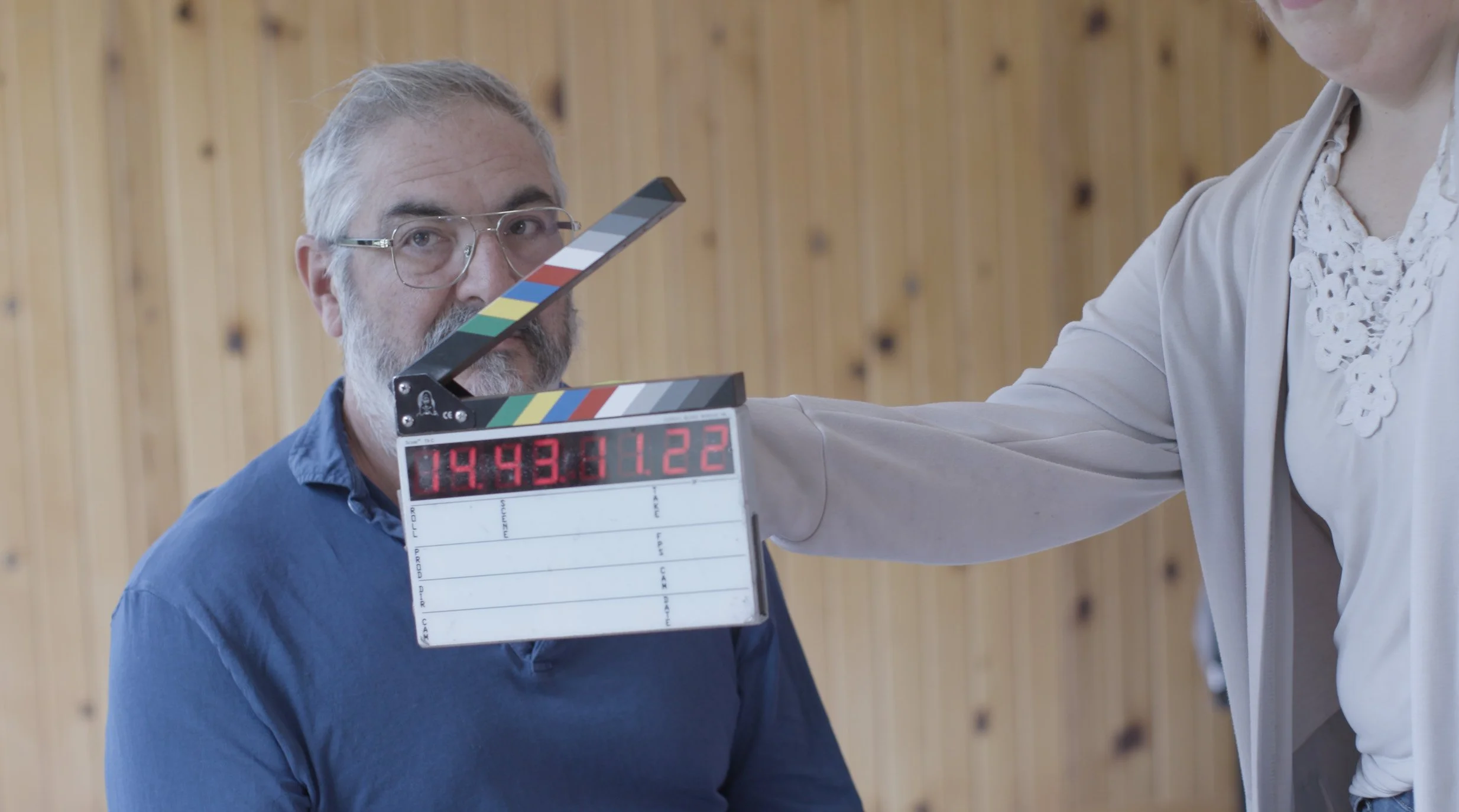 A man with gray hair and glasses sitting in front of a wooden wall, with a woman in a white blouse and beige jacket holding a clapperboard near his face.