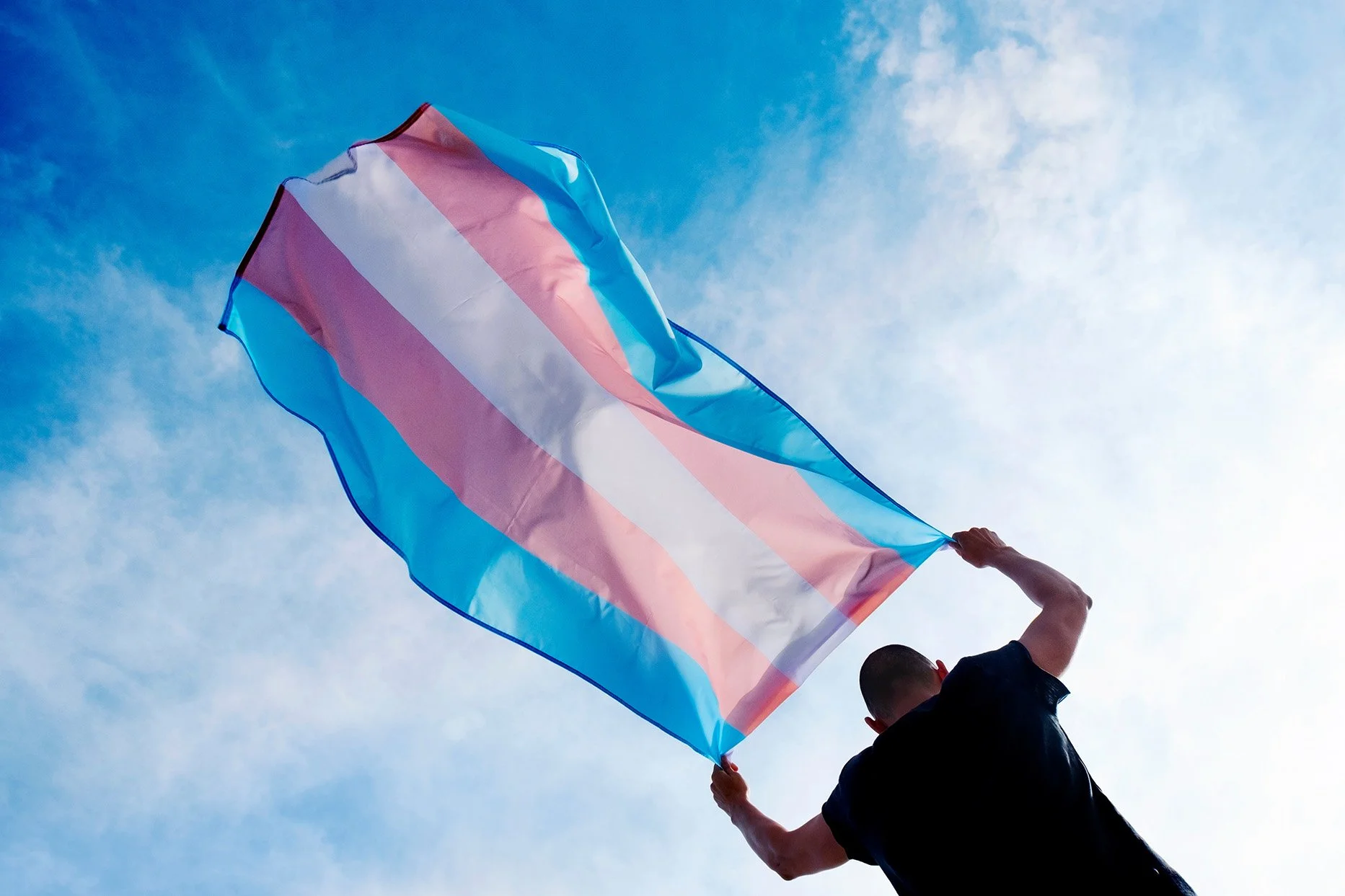 Person holding a transgender pride flag against a blue sky with some clouds.