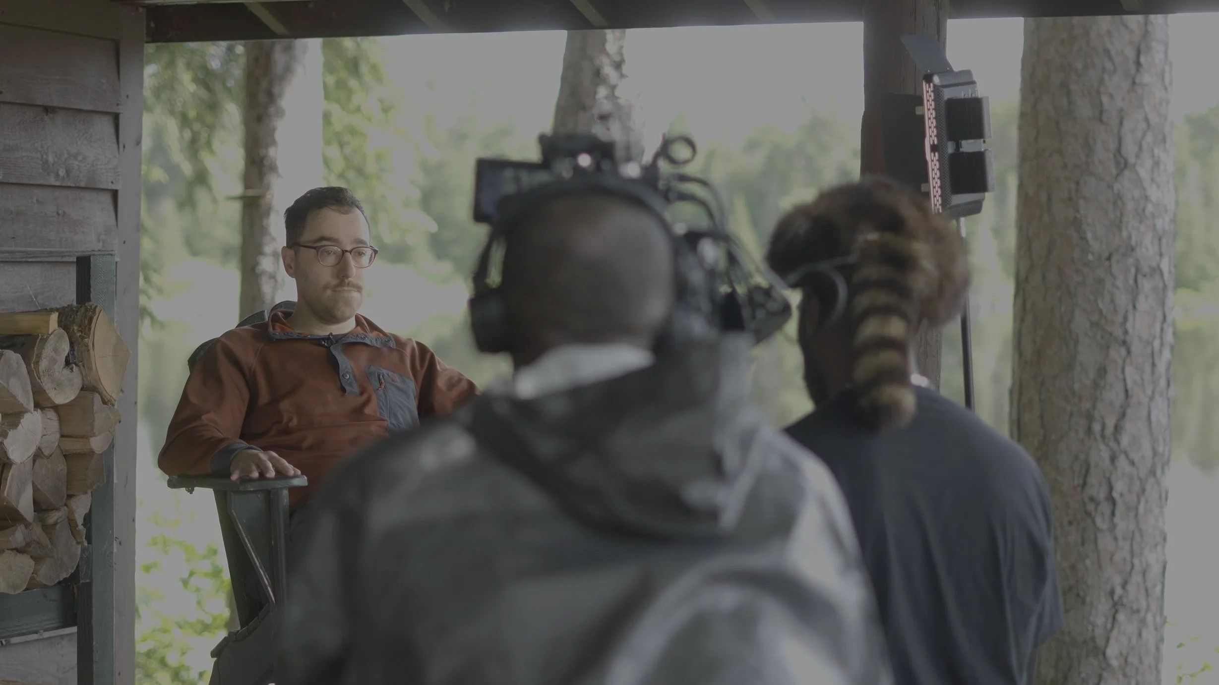 Man with glasses sitting on a chair during an outdoor interview, with two crew members filming him; one crew member has a raccoon resting on their shoulder.