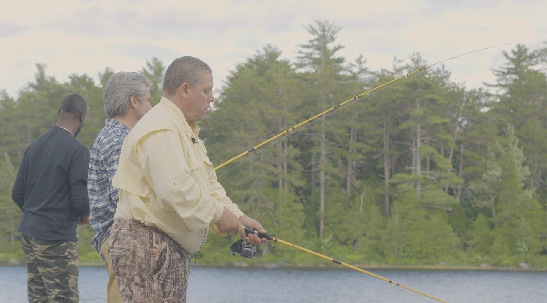 Three men fishing off a lakeshore with trees in the background.