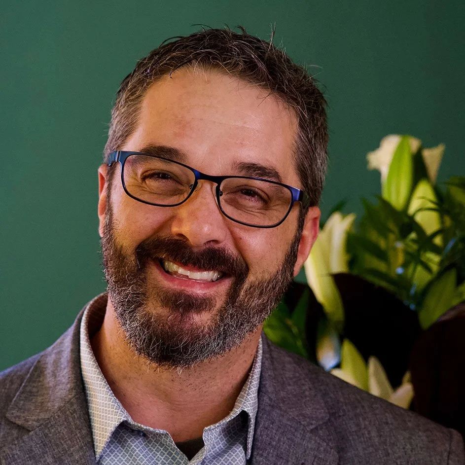 A man with short dark hair, beard, and mustache, smiling, wearing a colorful plaid shirt, standing against a plain white wall.