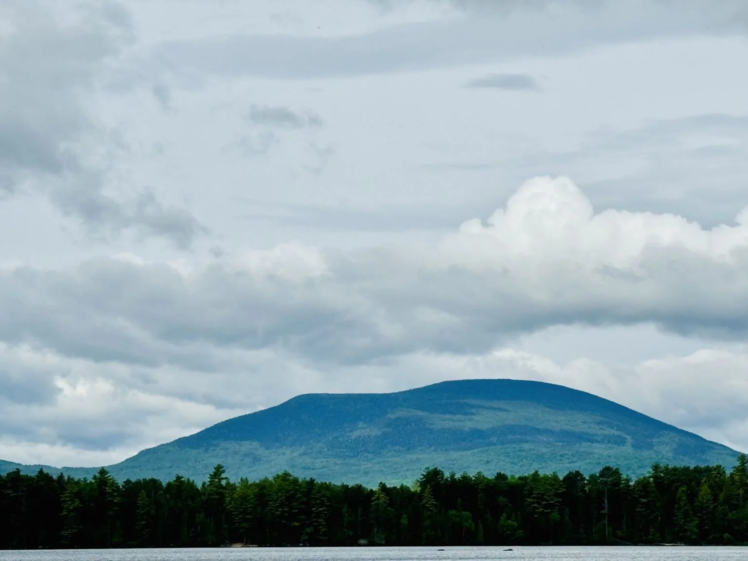 A landscape with a mountain in the background, a forest at the base, and a body of water in the foreground, under a cloudy sky.