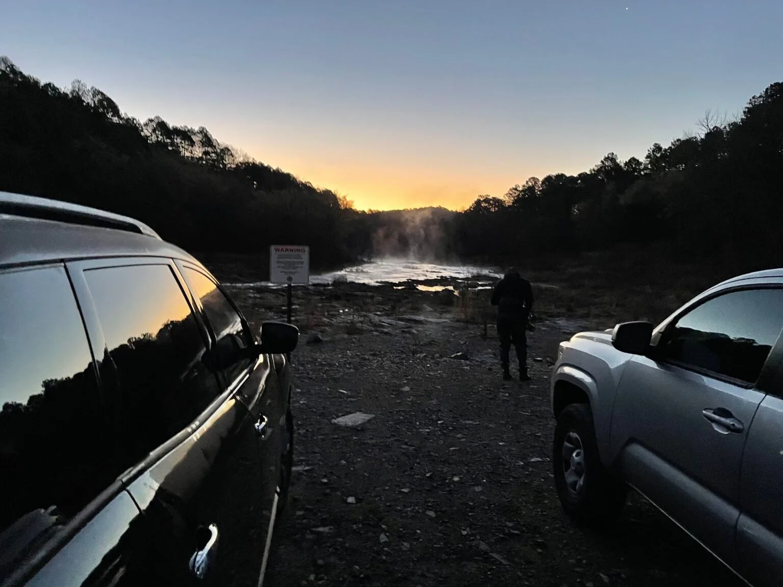 Two cars parked on a rocky terrain near a river at sunset, with a person standing near the river and a warning sign