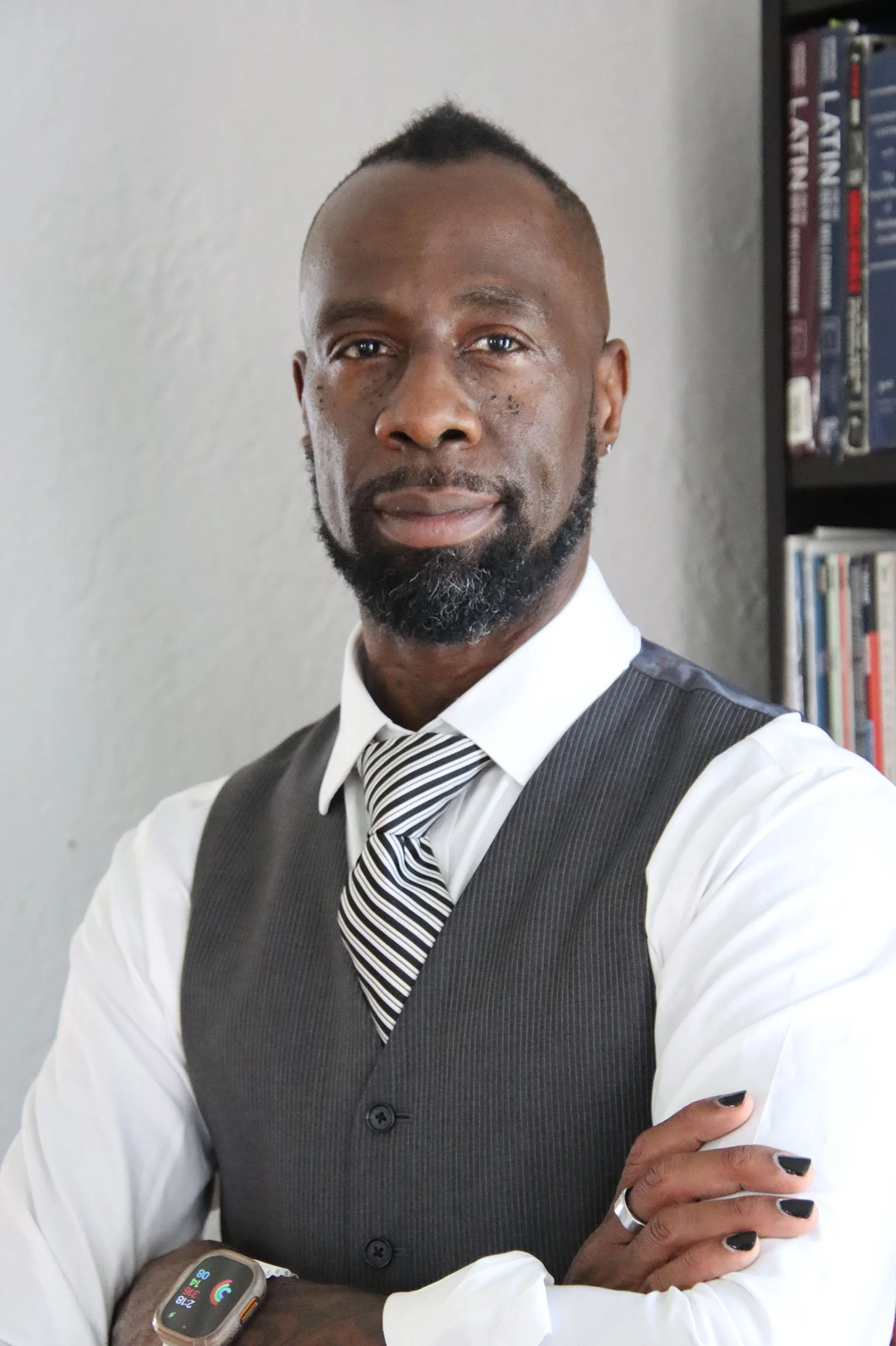 A professional Black man in a white shirt, striped tie, and gray vest standing with arms crossed in front of a bookshelf.