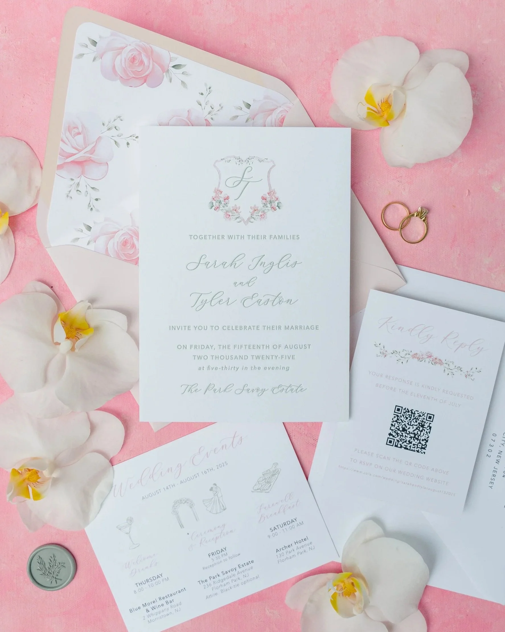 Wedding invitation suite with floral accents on pink background, surrounded by white orchids, a wedding ring, and a coin.