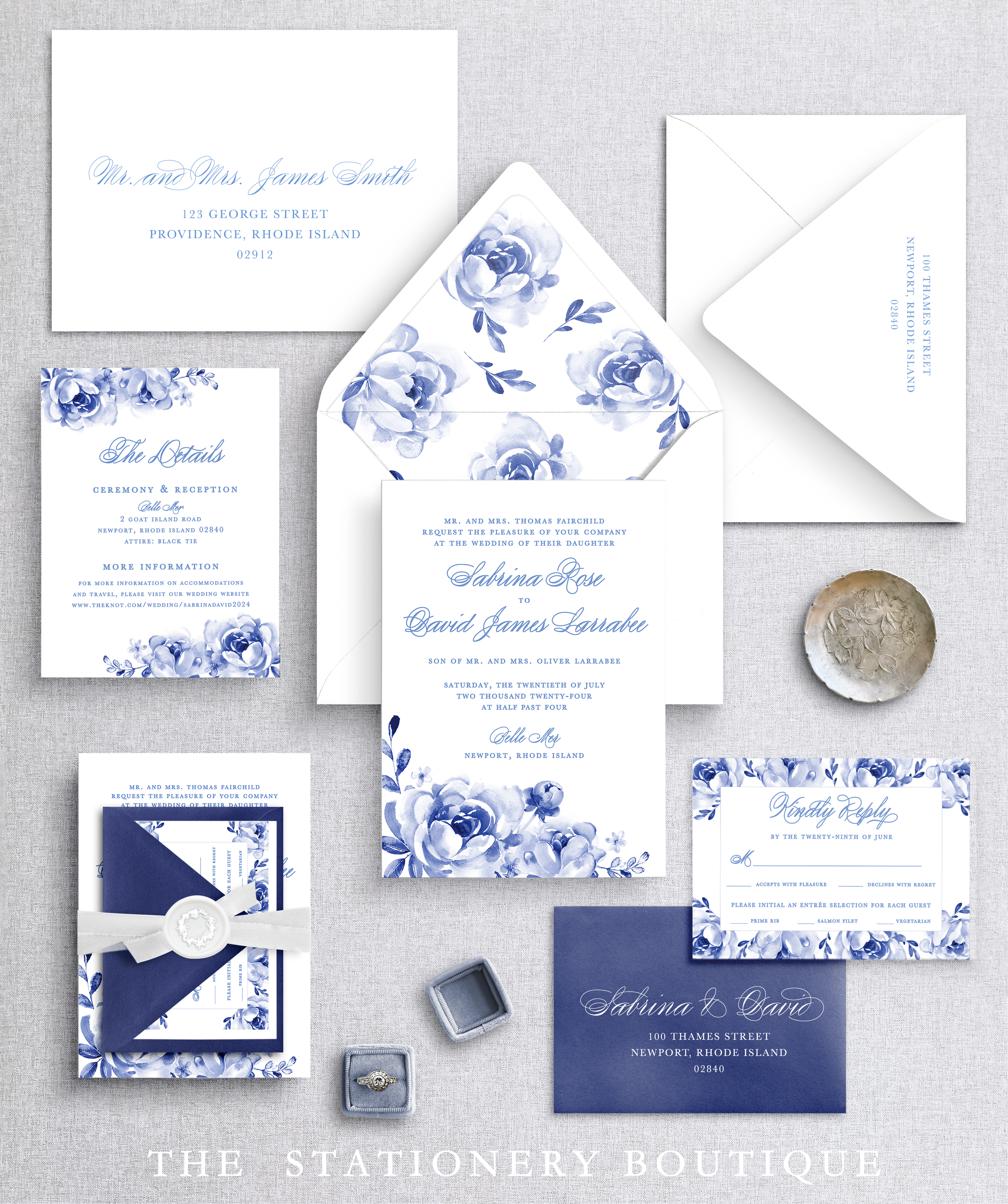 Flat lay of elegant wedding stationery with blue floral designs, including invitations, reply card, envelopes, and ring boxes on a gray fabric background.