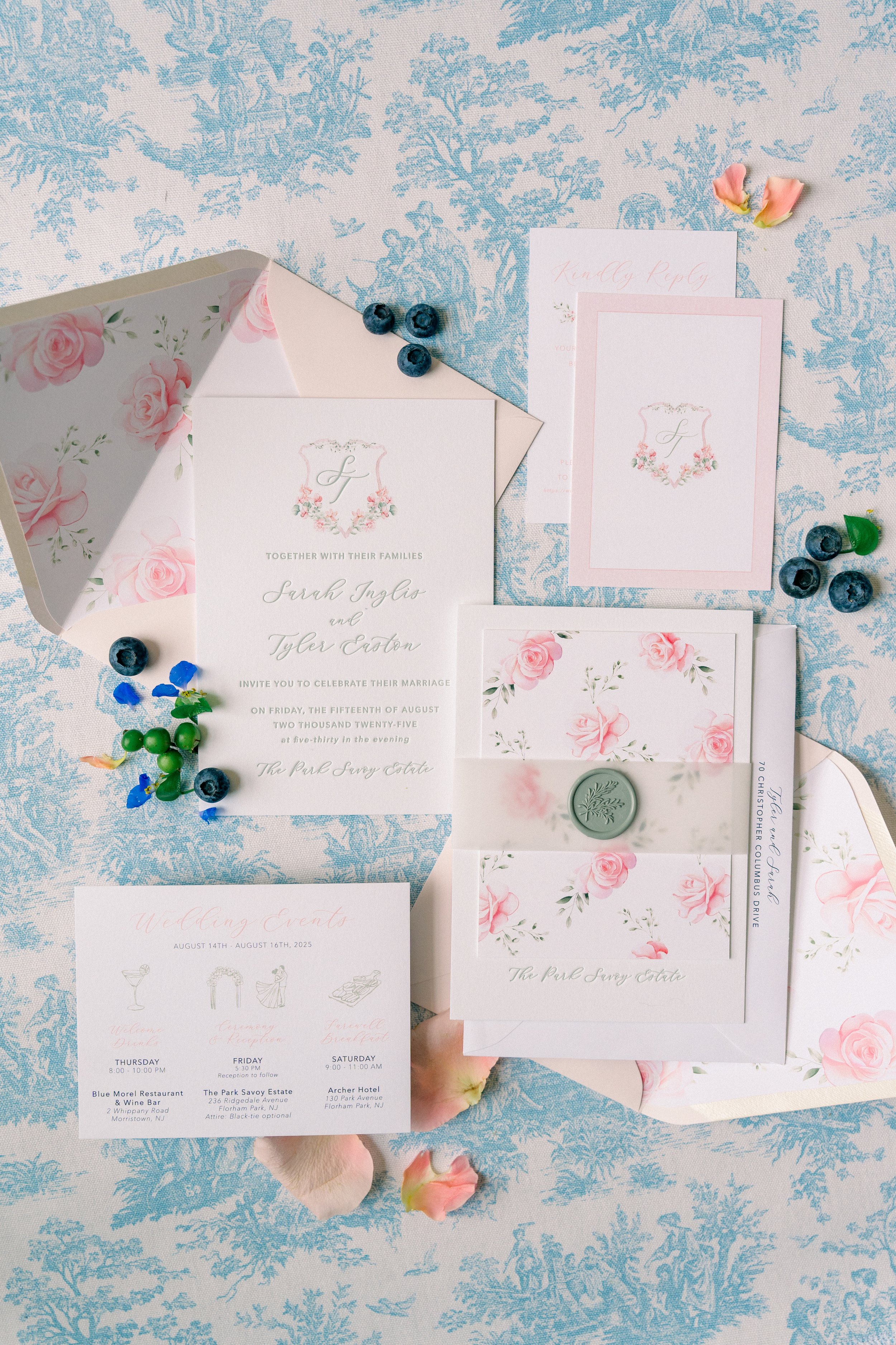 Wedding invitation suite with pink roses and greenery, blue berries, and scattered flower petals on a blue and white patterned tablecloth.