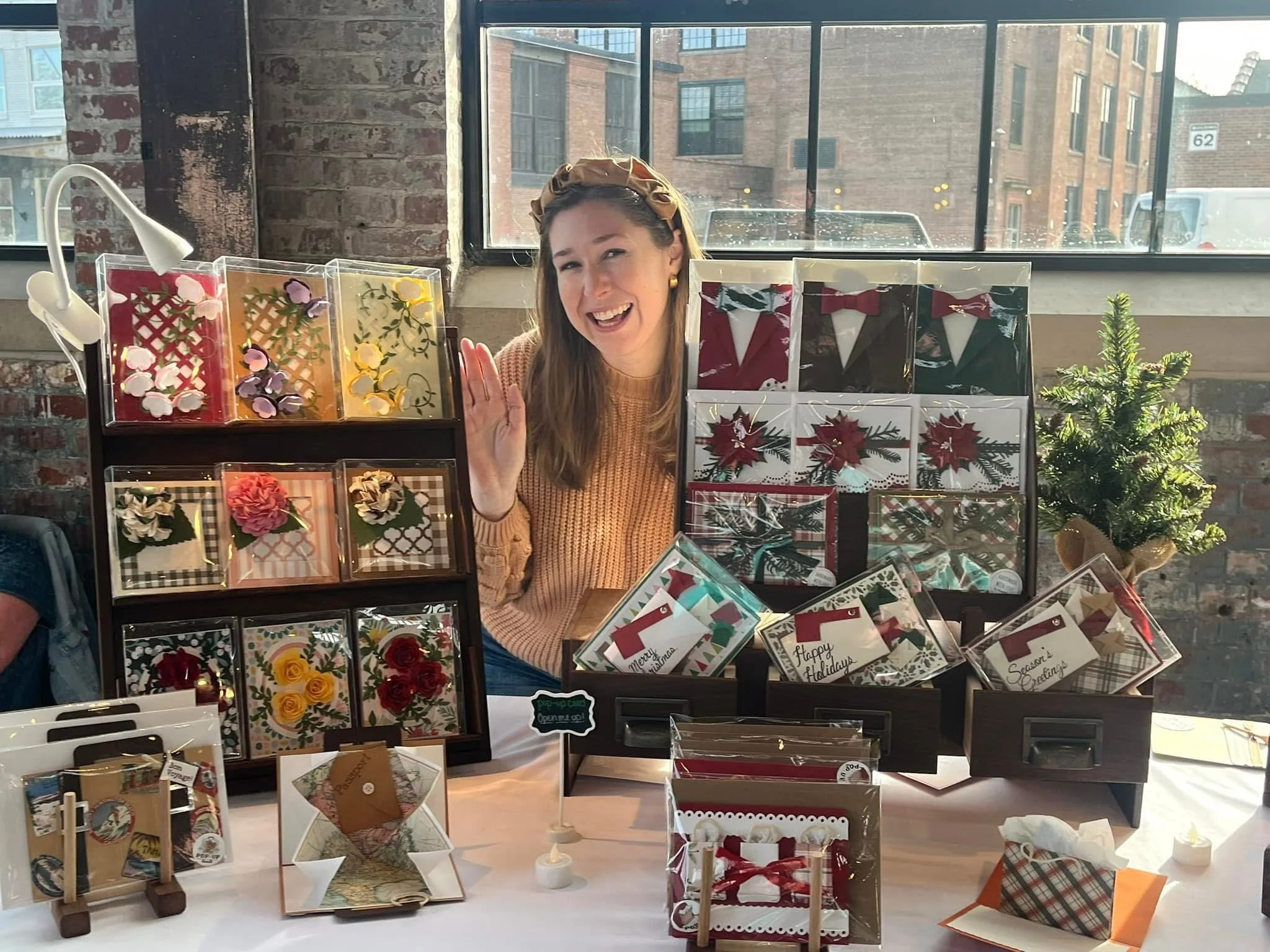 A woman at a Christmas craft stall with holiday-themed decorations and greeting cards, smiling and waving.