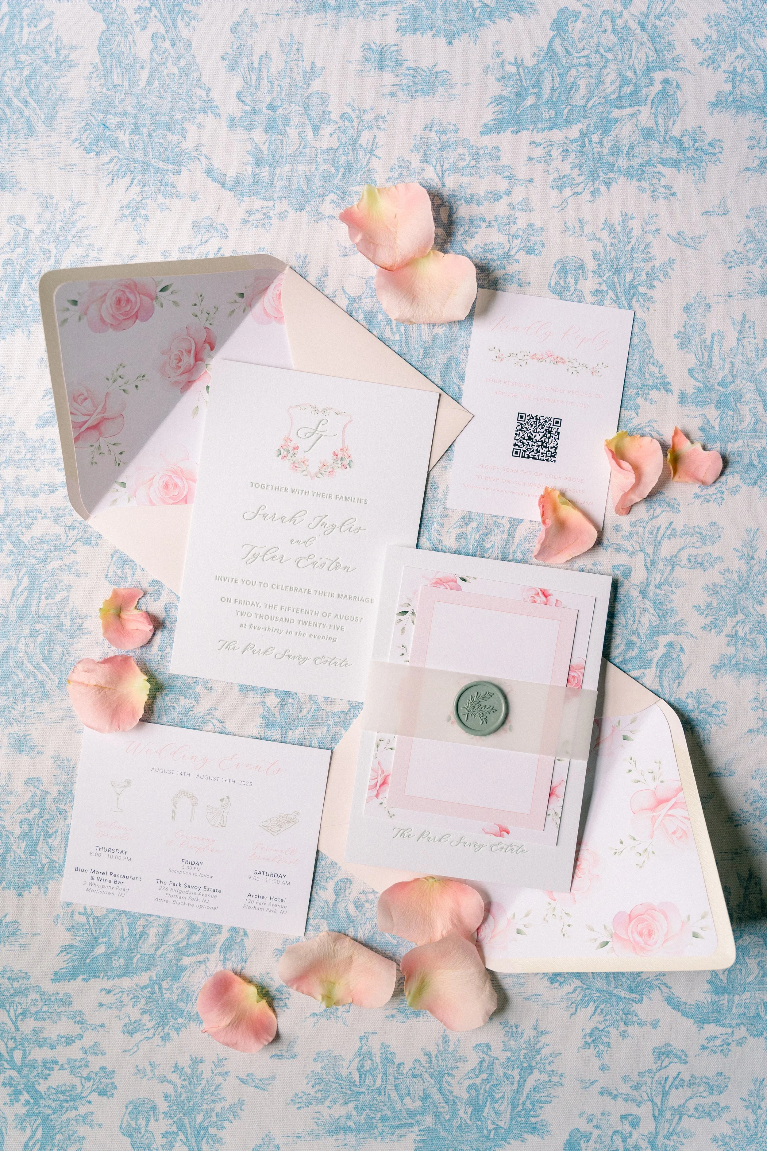 A wedding invitation set with floral designs, a sealed envelope, a pink wax seal, an RSVP card with a QR code, and a wedding schedule, all arranged on a light blue and white patterned fabric with pink rose petals scattered around.