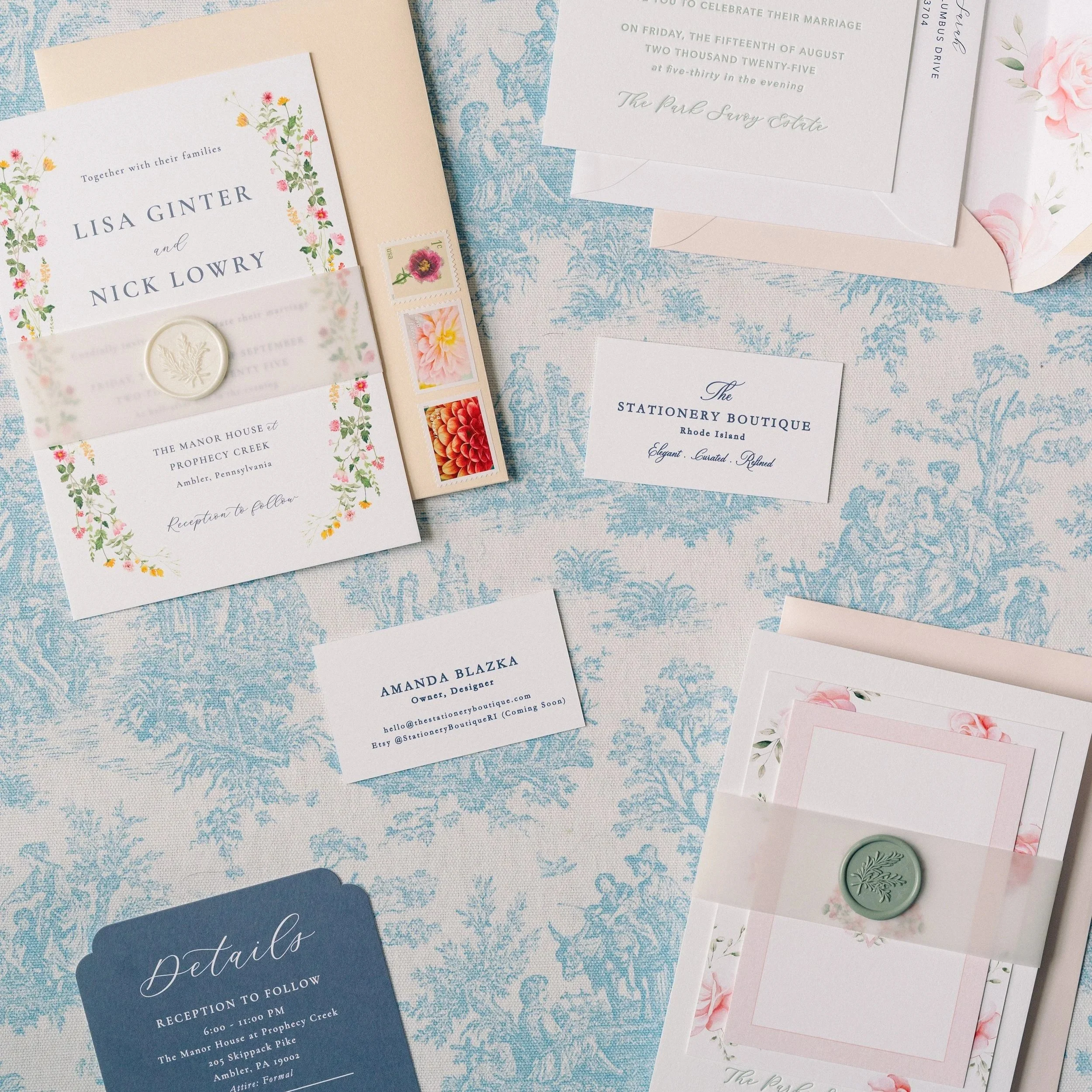 Wedding invitation suite with floral details, place cards, and postage stamps on a blue and white patterned tablecloth.