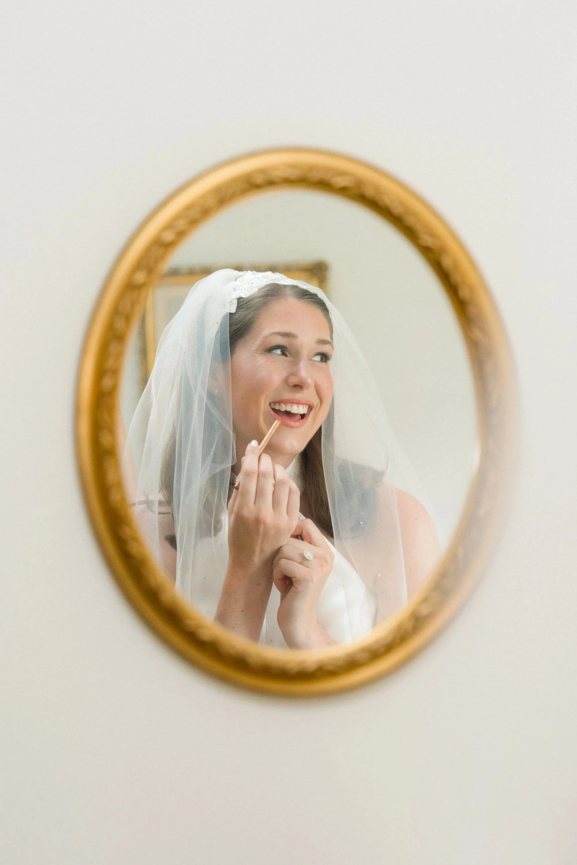 A woman in a wedding dress and veil applying lipstick while looking in a mirror.
