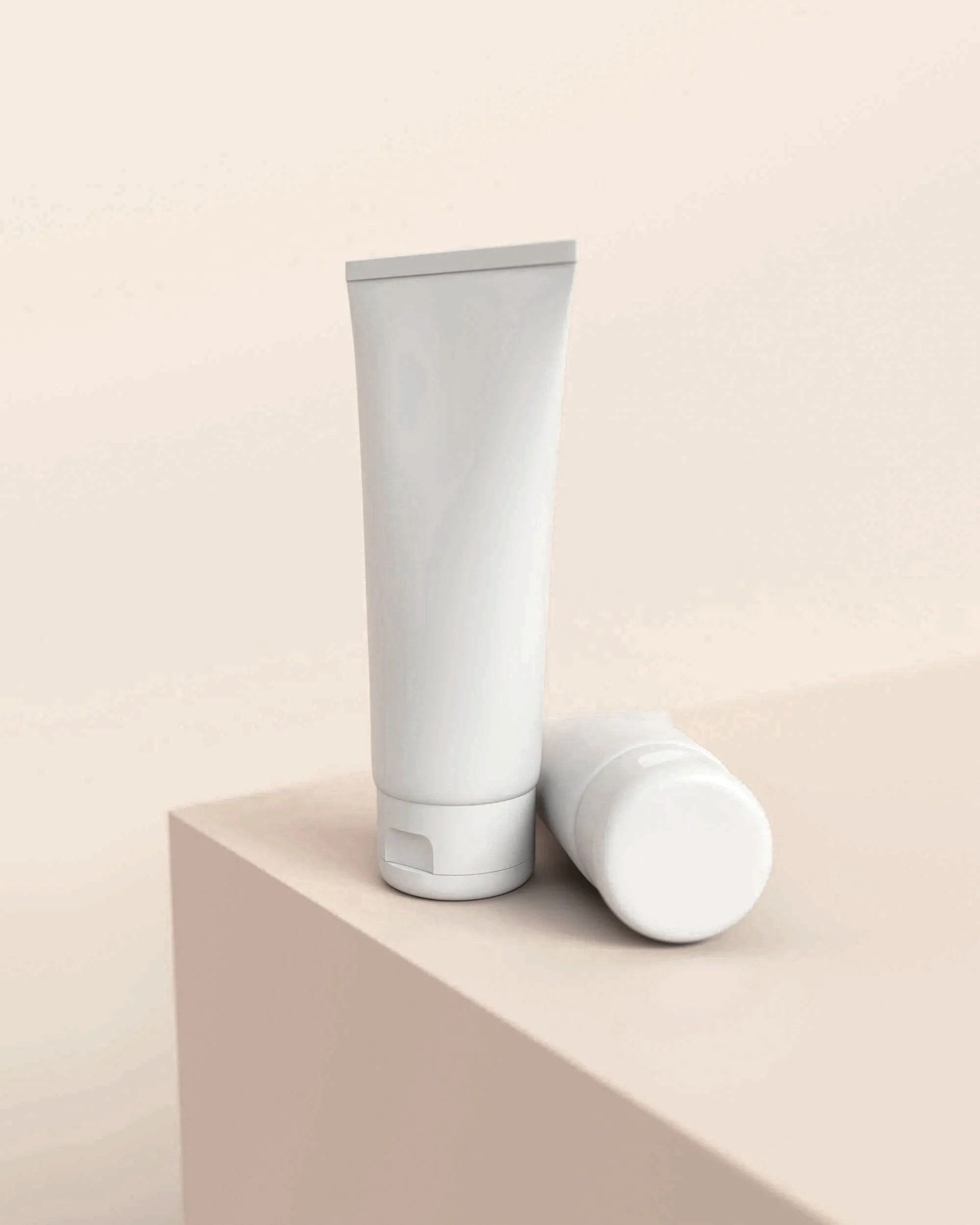 Two white cosmetic tubes, one standing upright and one lying down, on a light-colored surface against a neutral background.
