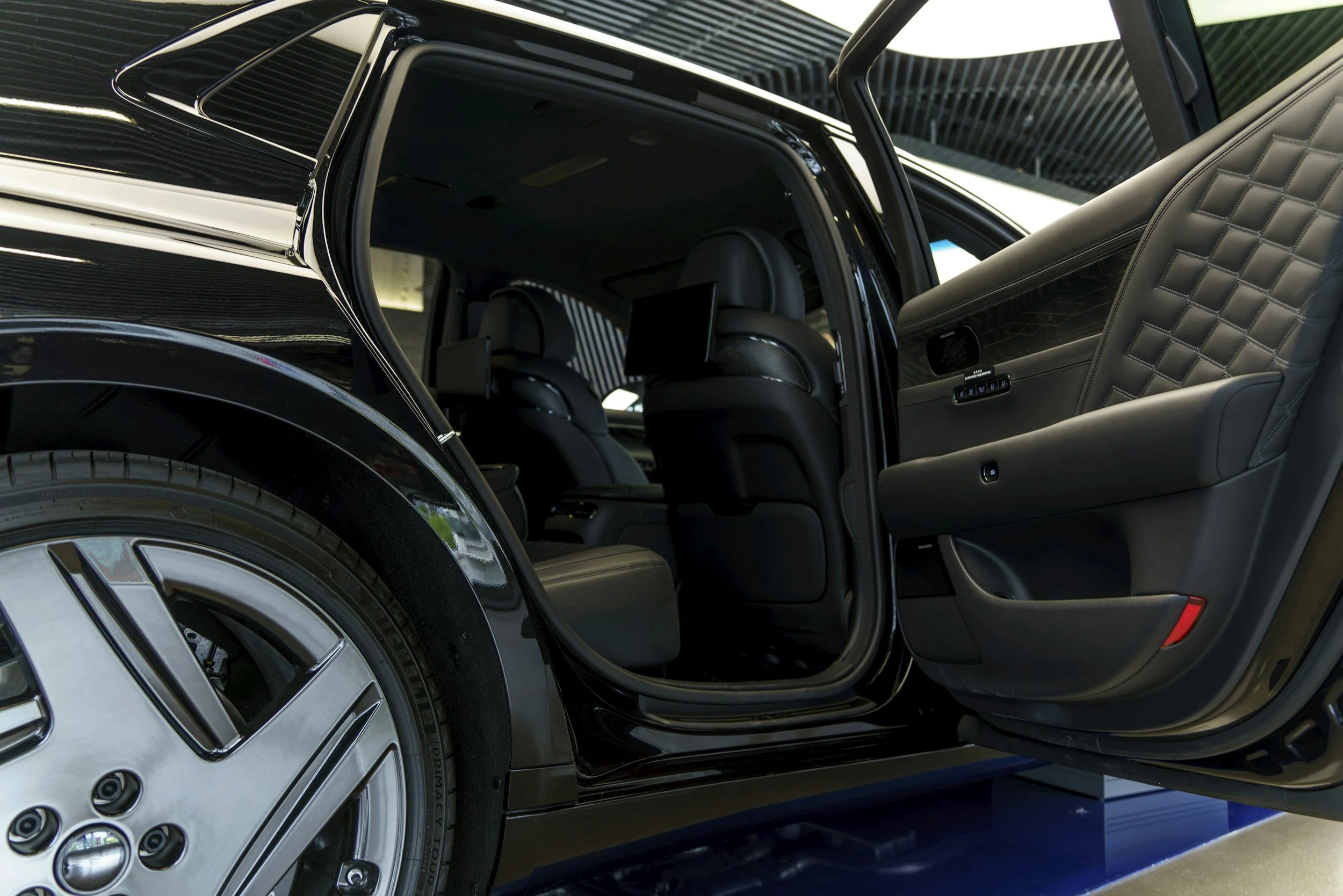 Close-up of a luxury black car interior with open rear door, showing black leather seats and a quilted door panel, with part of the tire and wheel visible.