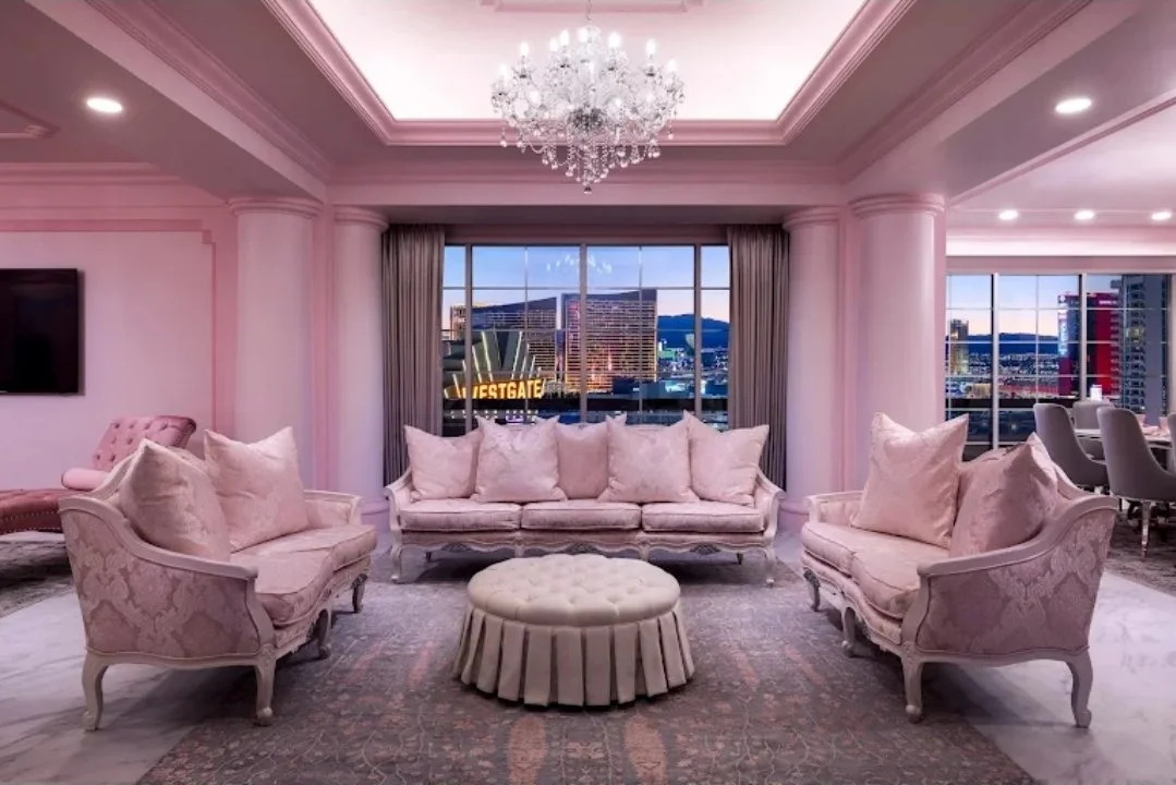 Elegant pink living room with ornate furniture, large windows showing cityscape, and a chandelier.