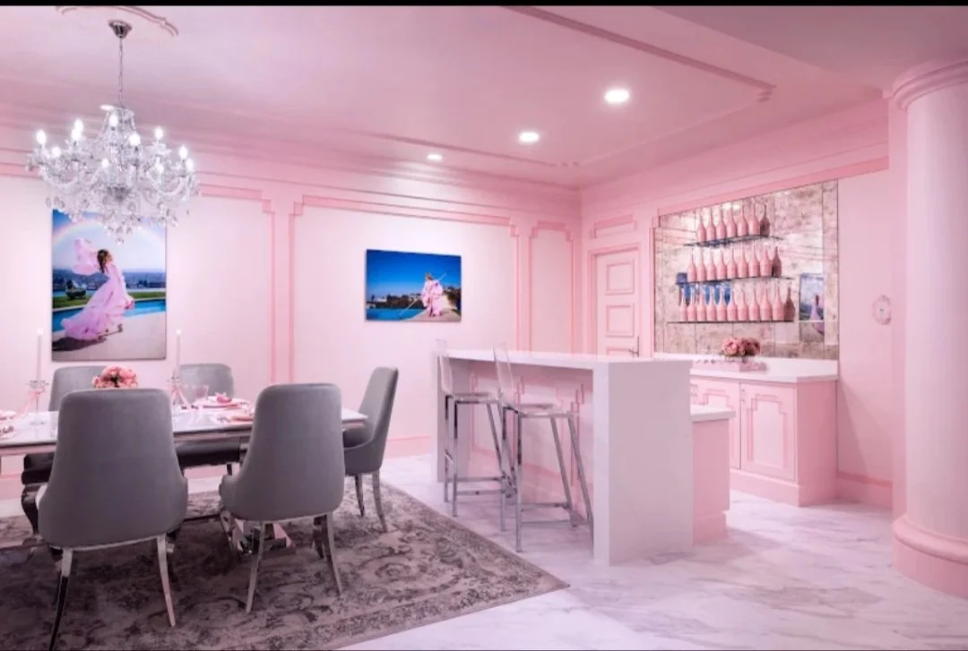 A pink-themed dining area with a chandelier, a dining table with six chairs, and a bar area with barstools. Decor includes framed photographs and vases with pink flowers.