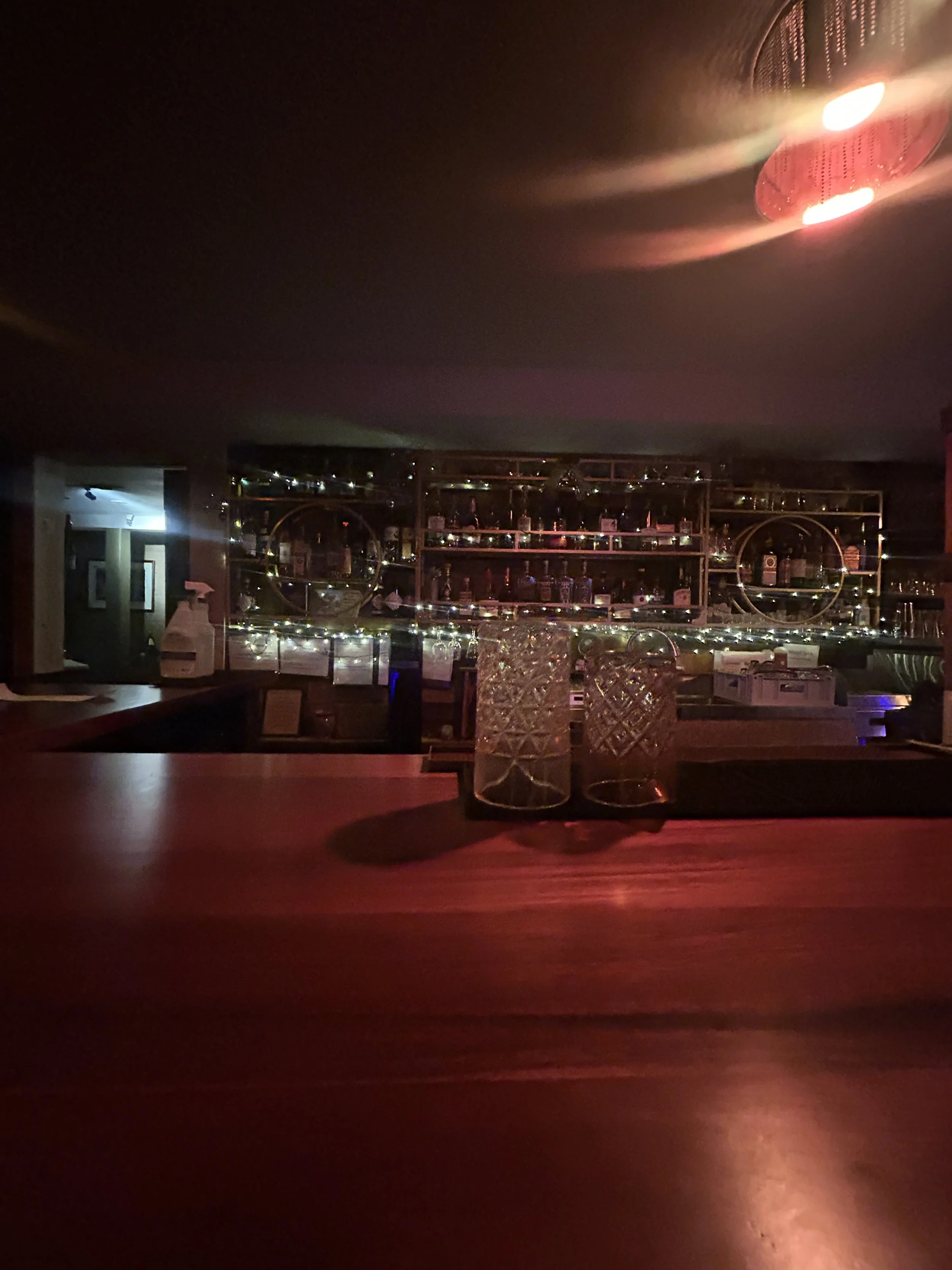 Dimly lit bar with glassware and bottles on shelves, illuminated by decorative string lights, with two glasses on the bar counter.
