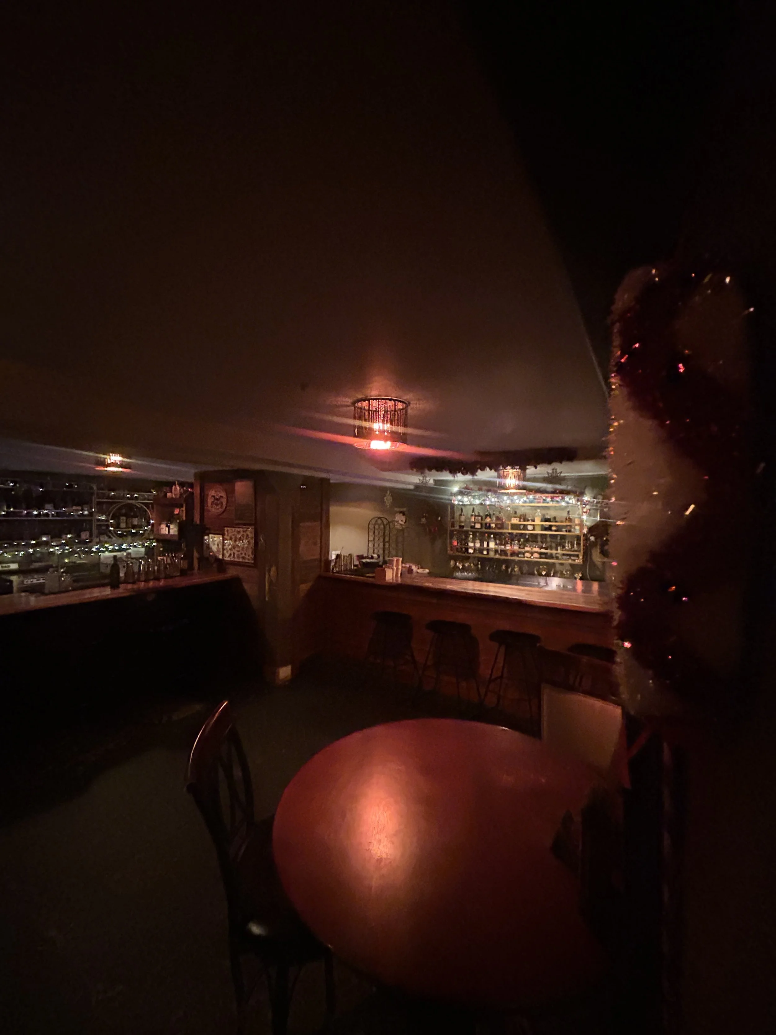 Dimly lit bar with a round table and chairs, bar counter with stools, shelves stocked with bottles, and Christmas decorations. Red ceiling lights create a cozy atmosphere.